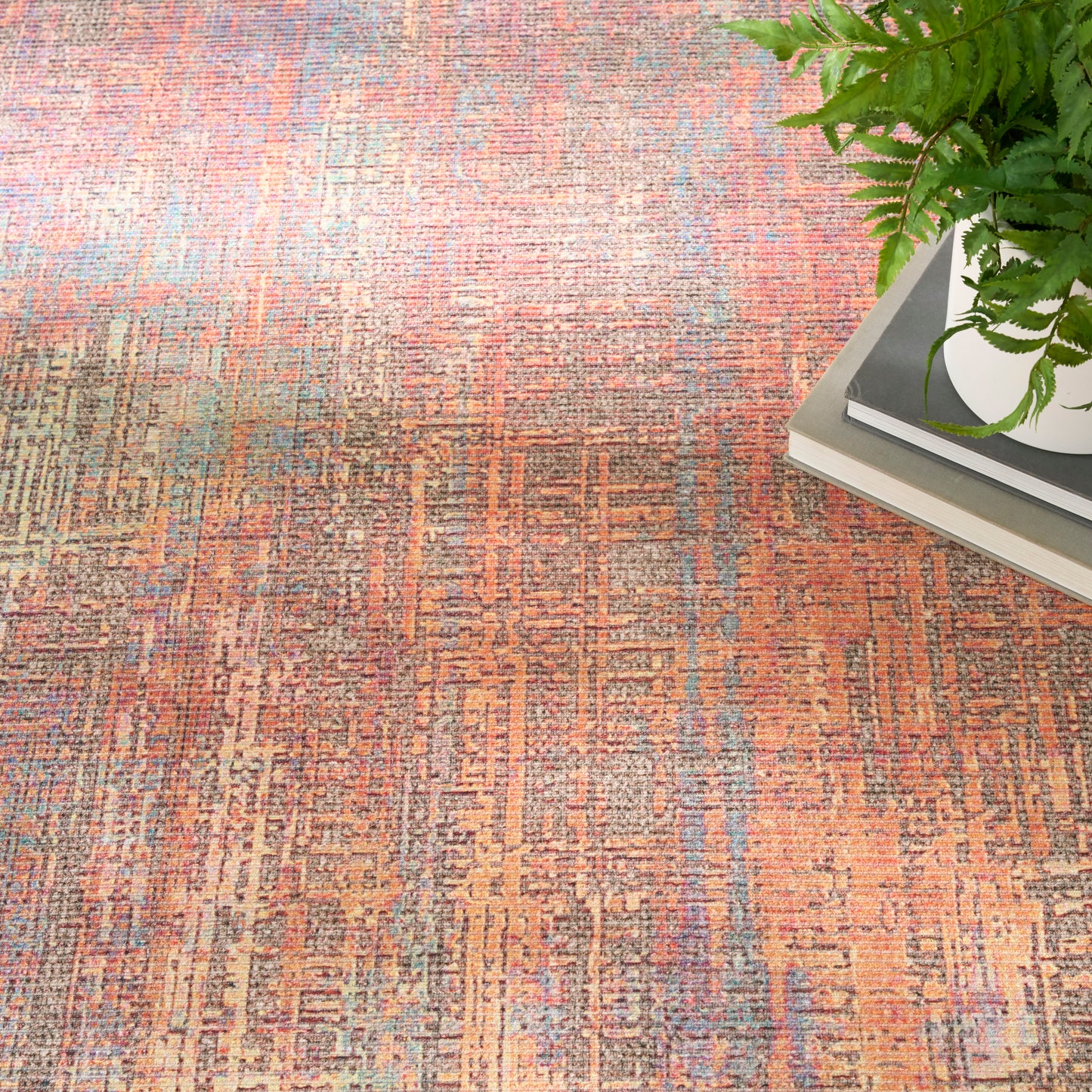 Orange-brown geometric patterned area rug with teal accents, beside a plant on a stack of books.