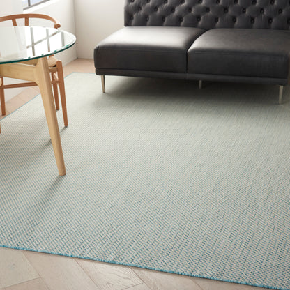 Pale textured rug (8' x 10') in a living room setting, with a dark gray tufted leather sofa on slender metal legs beside a glass round table.