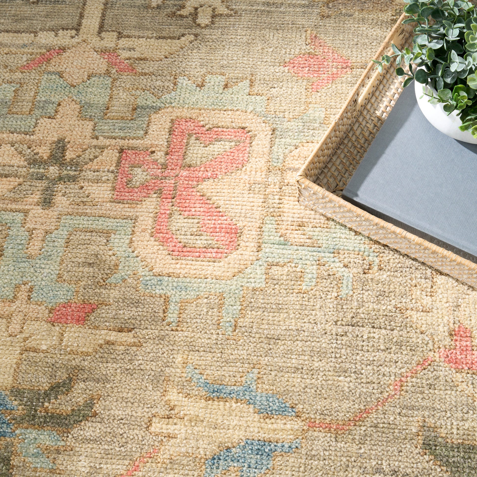 Beige woven rug with pink, blue and green geometric motifs; textured surface; plant in a white pot on a wicker tray nearby.