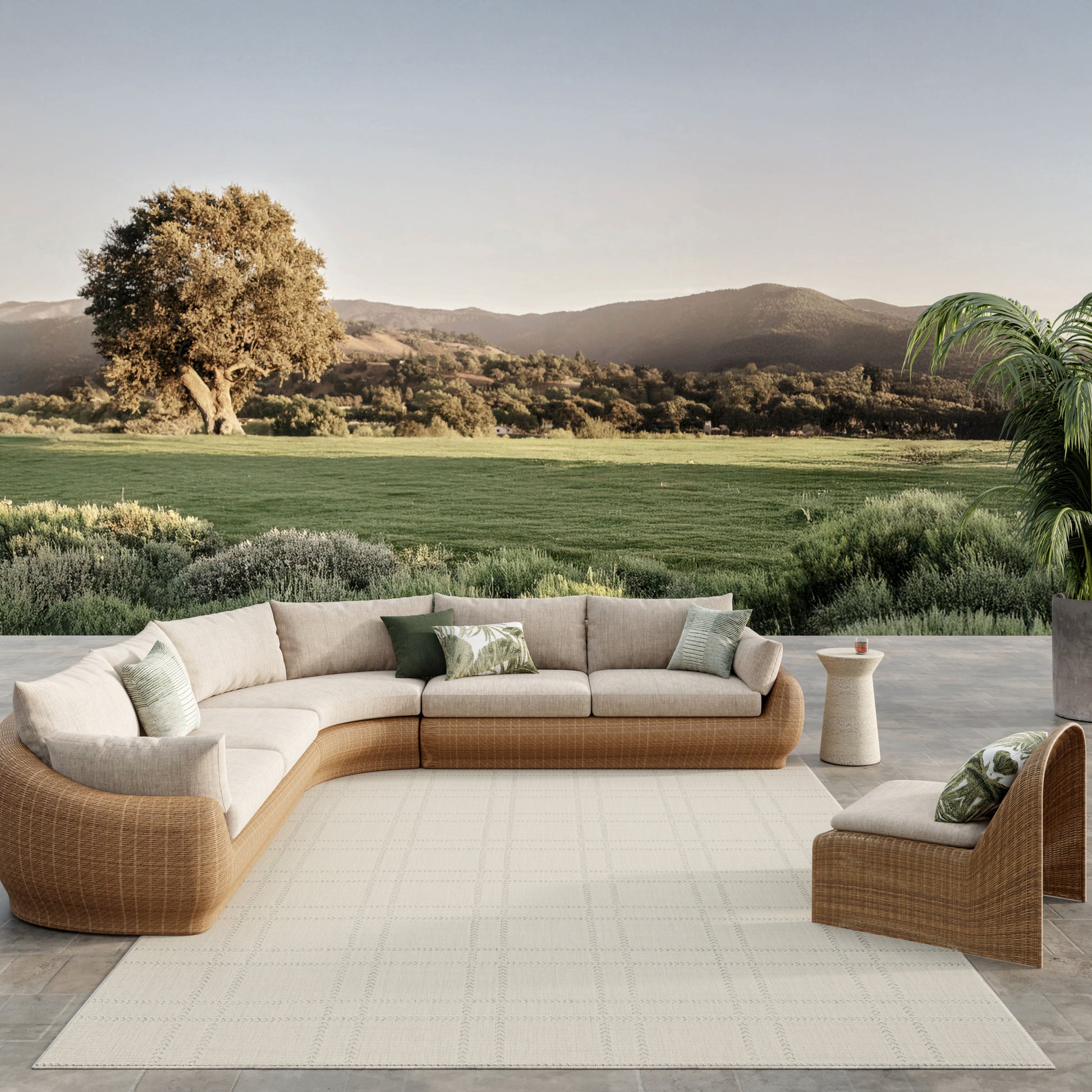 Beige cushioned rattan outdoor sectional with a matching chair on a cream rug, round side table, set in a scenic countryside.