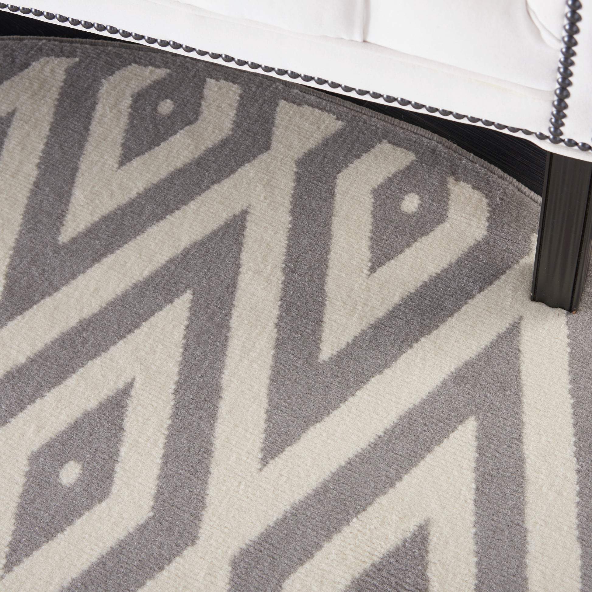 Gray & cream chevron geometric round rug under a white upholstered bed frame with nailhead trim & black leg.