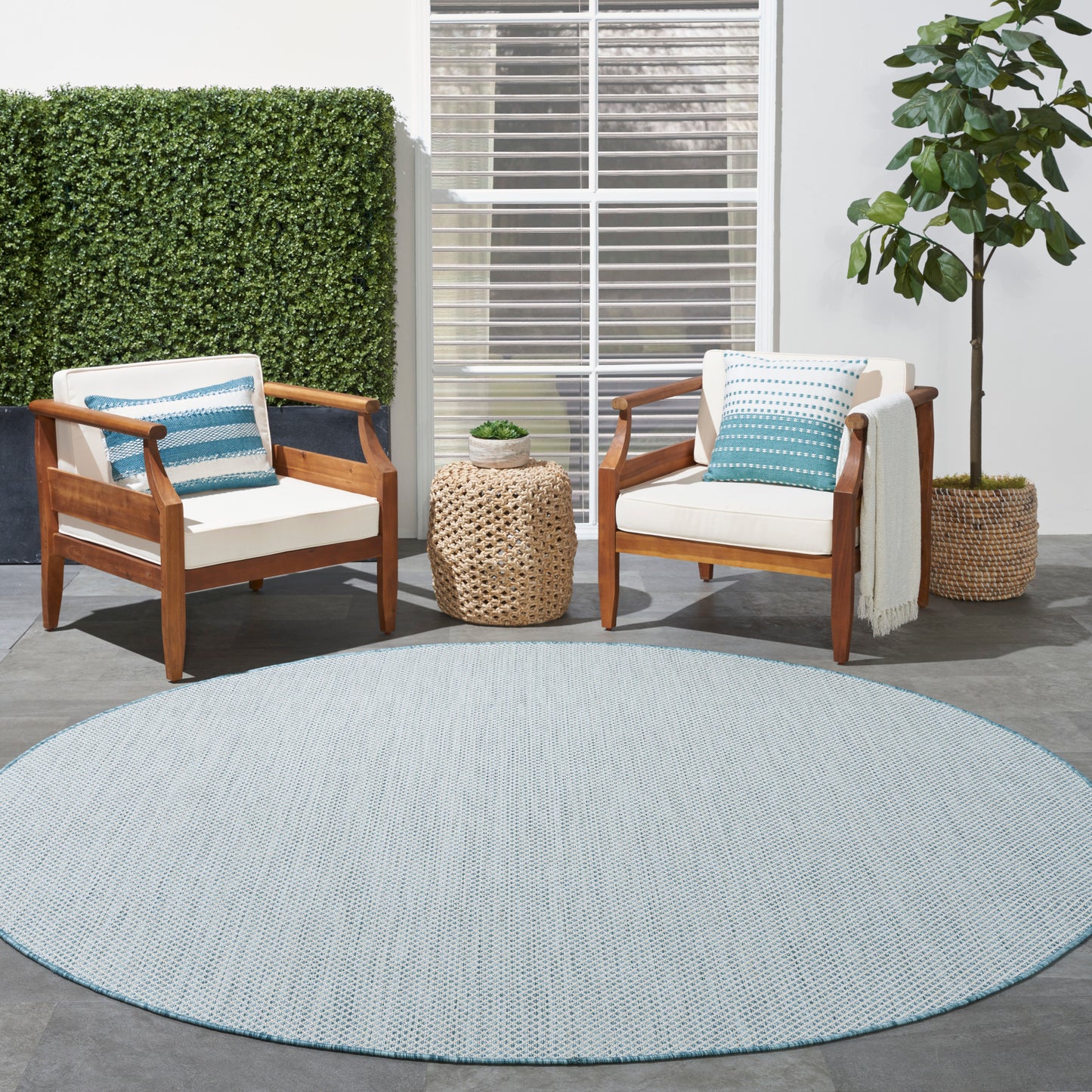 Outdoor seating: two wooden armchairs with cream cushions and blue pillows, round blue rug, wicker side table, potted plant.