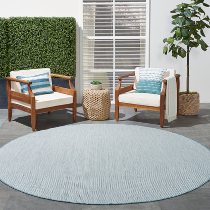 Outdoor seating: two wooden armchairs with cream cushions and blue pillows, round blue rug, wicker side table, potted plant.