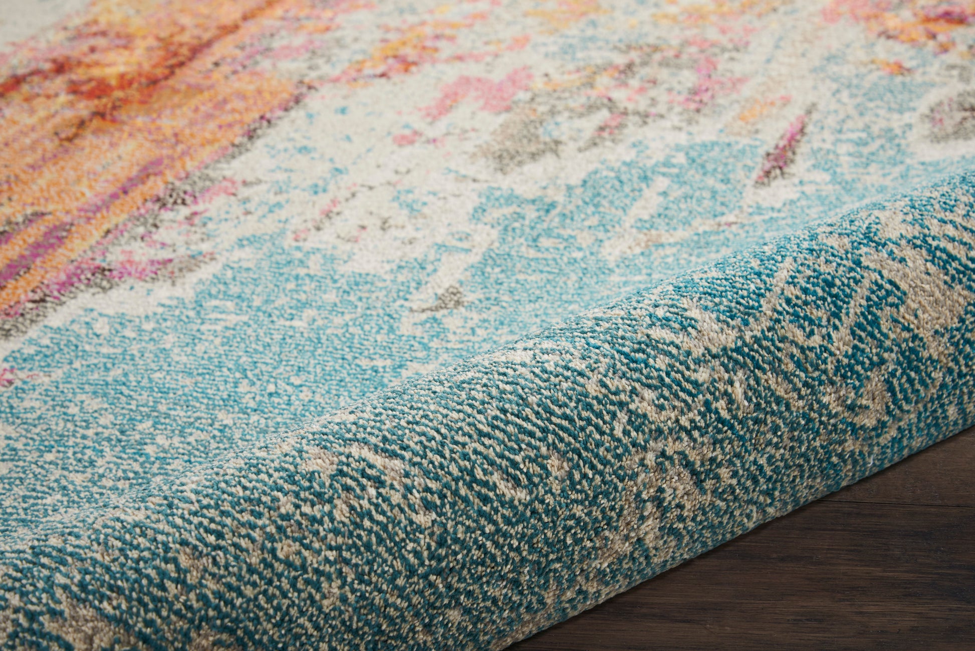 Close-up of a multicolor woven area rug with teal border & abstract pink, orange, blue pattern on a wooden floor.