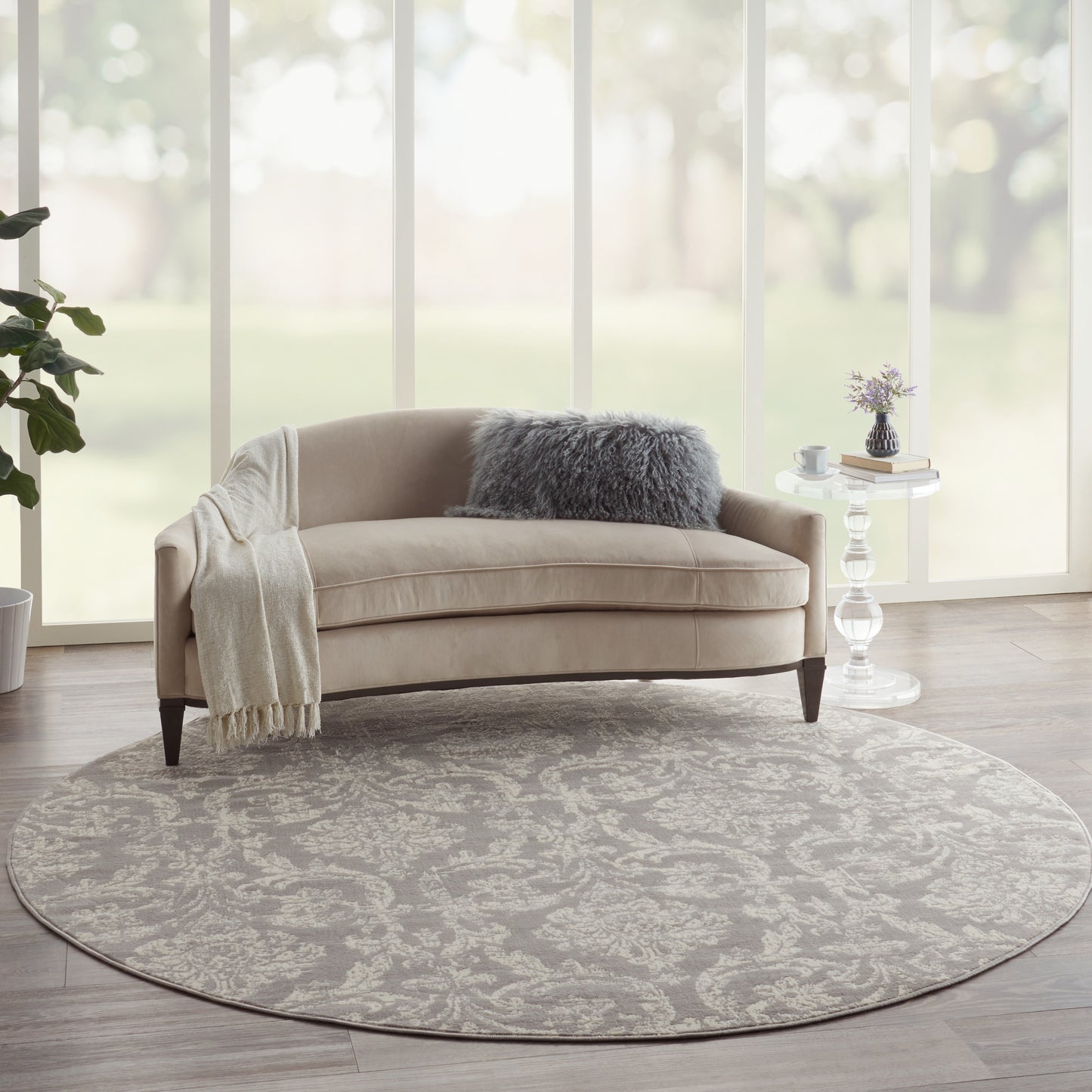 Beige curved sofa with cream throw and plush gray pillow on round grey damask rug; glass side table and greenery.