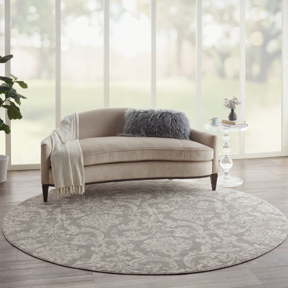 Beige curved sofa with cream throw and plush gray pillow on round grey damask rug; glass side table and greenery.