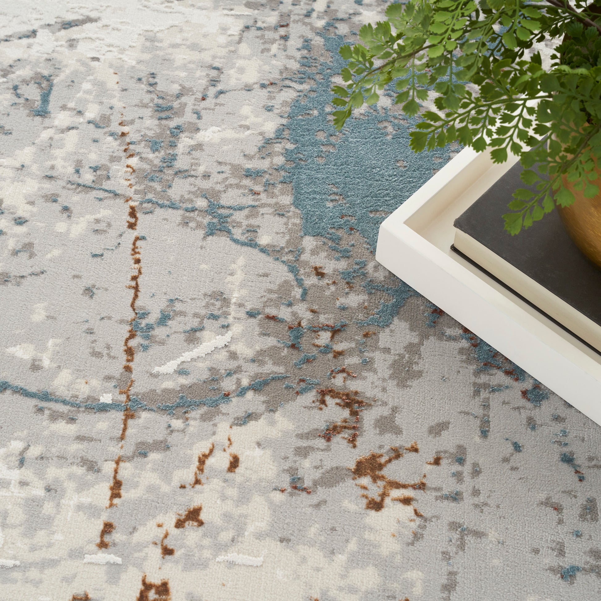 Abstract gray-blue rug with rust-brown accents; white tray, books, & a green fern in a golden pot.