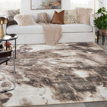 Living room scene with abstract brown-beige area rug, light gray sofa, textured throws, pillows, brass lamp, and plant.