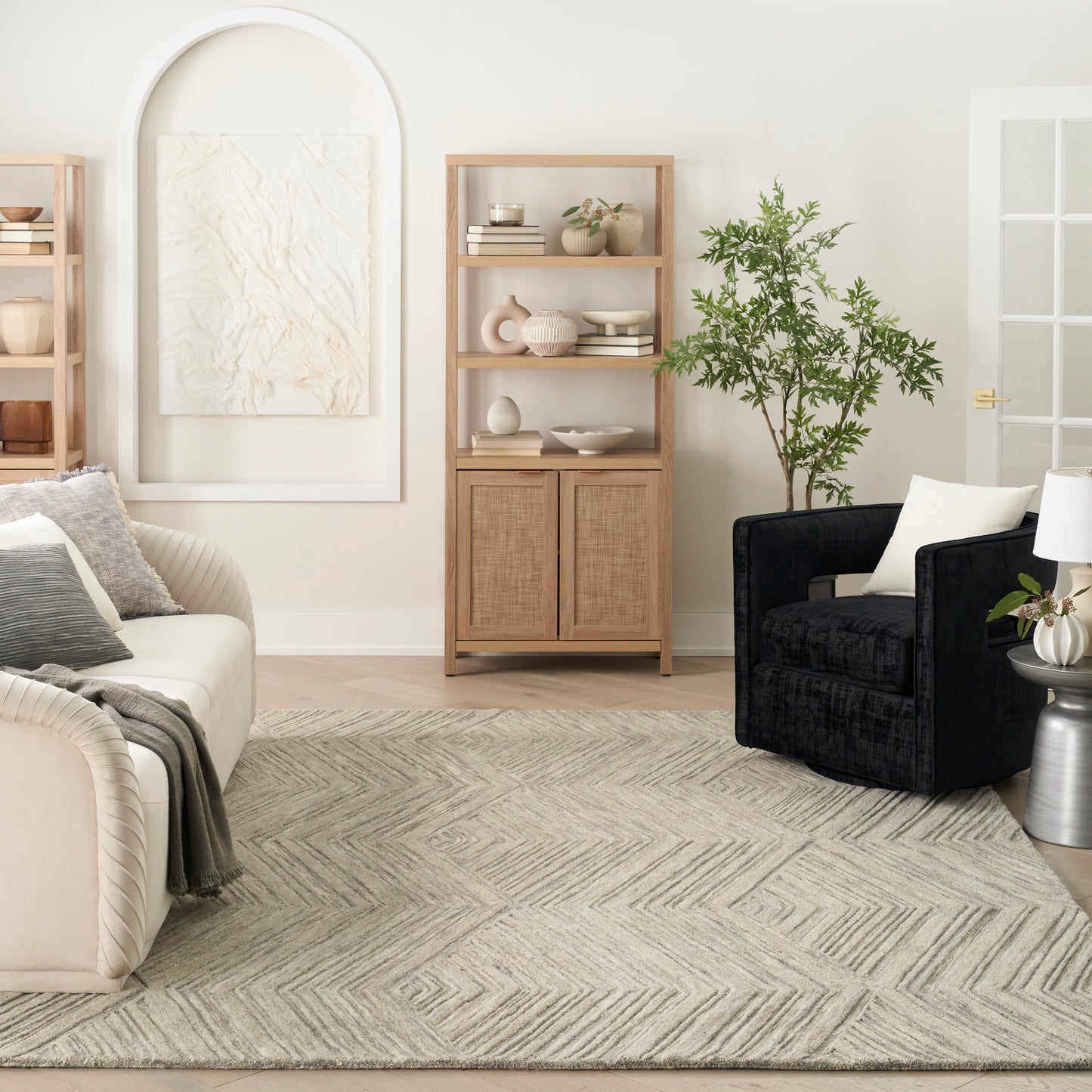 Geometric rug in a neutral living room with a beige sofa, black armchair, wooden shelving, arched panel art, and a leafy plant.