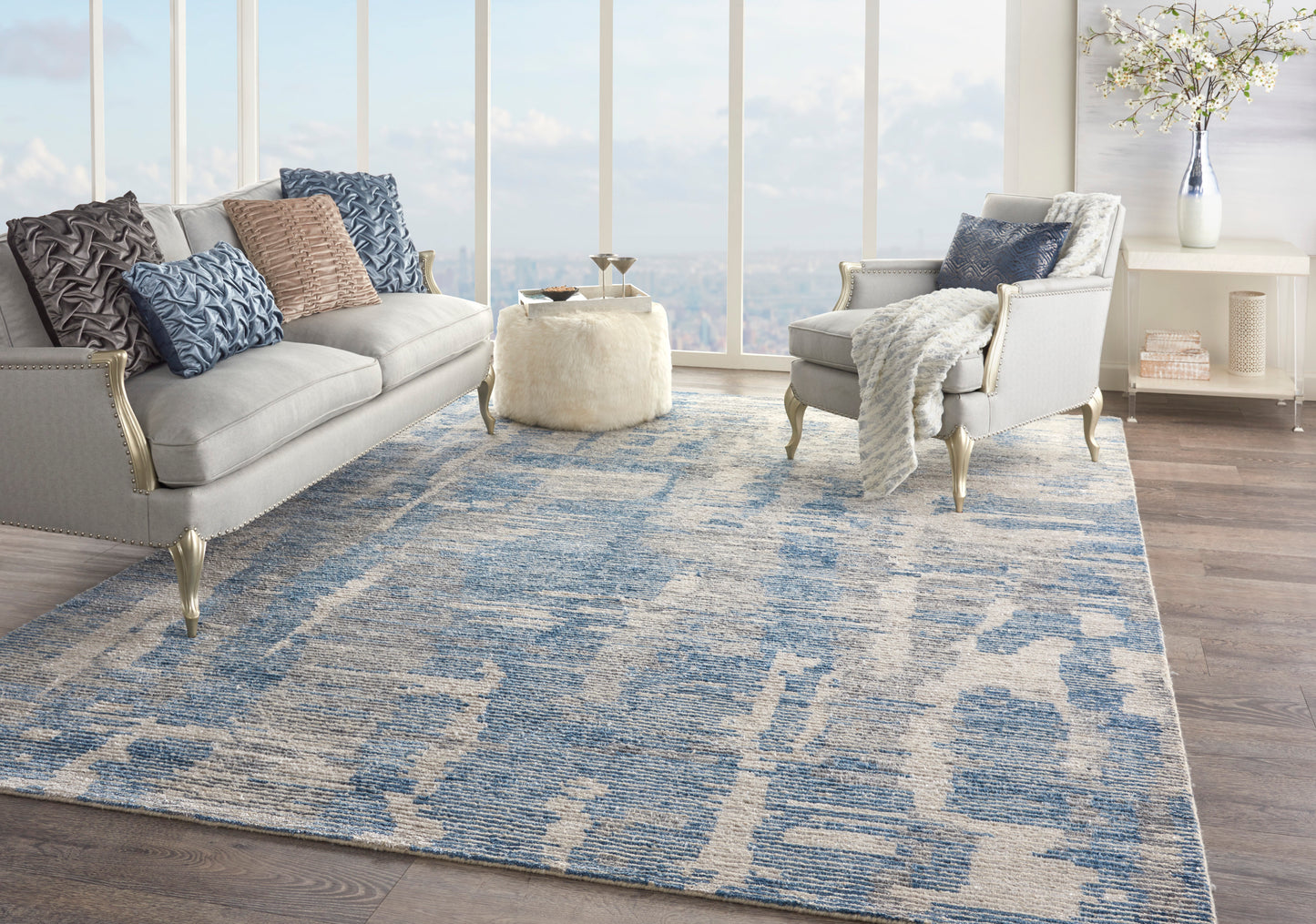 Living room with blue abstract rug, light gray sofas, blue/gray pillows, cream ottoman, white console, and large windows.