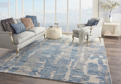 Living room with blue abstract rug, light gray sofas, blue/gray pillows, cream ottoman, white console, and large windows.