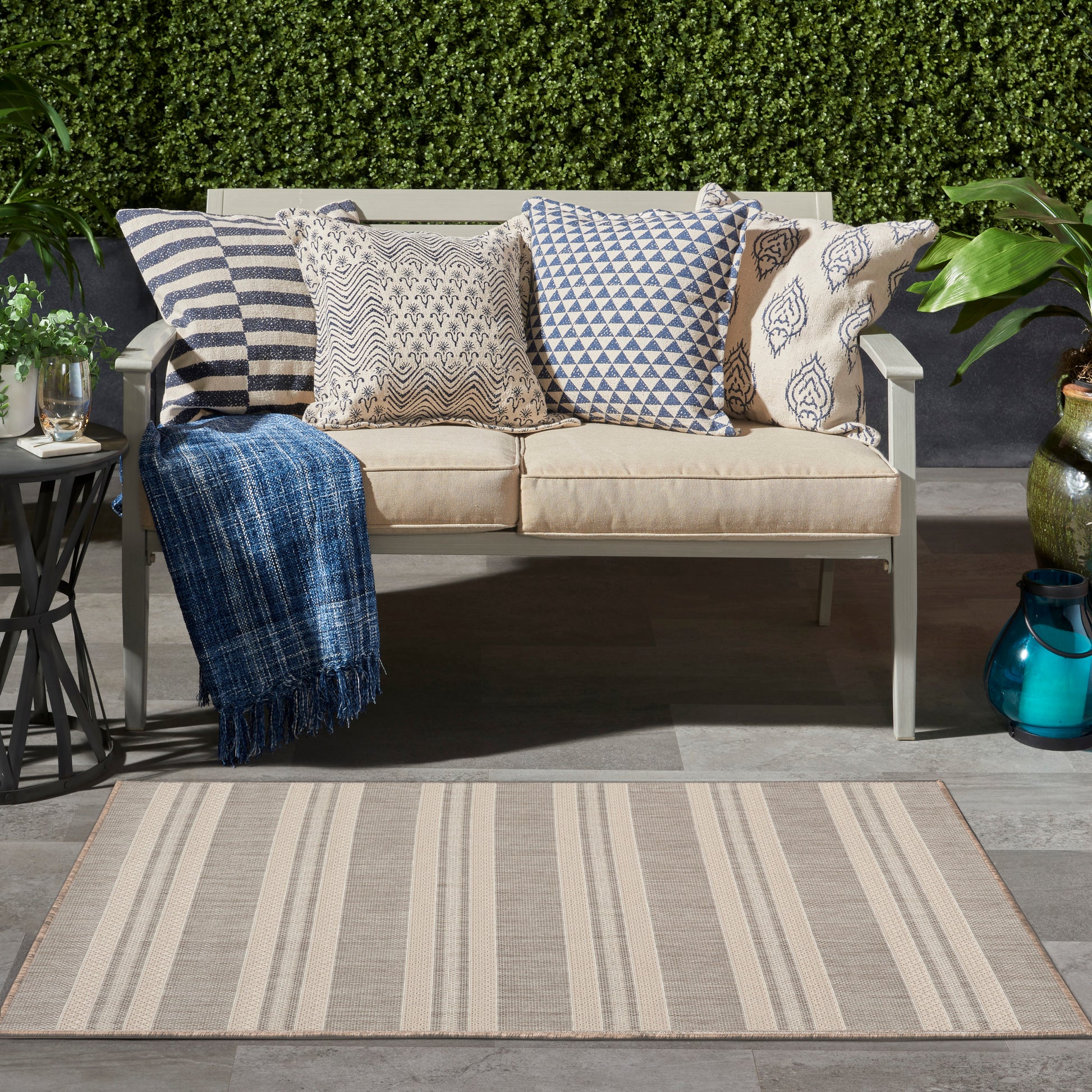 Beige outdoor loveseat with striped & geometric patterned cushions, blue throw, & beige striped rug against green hedge.