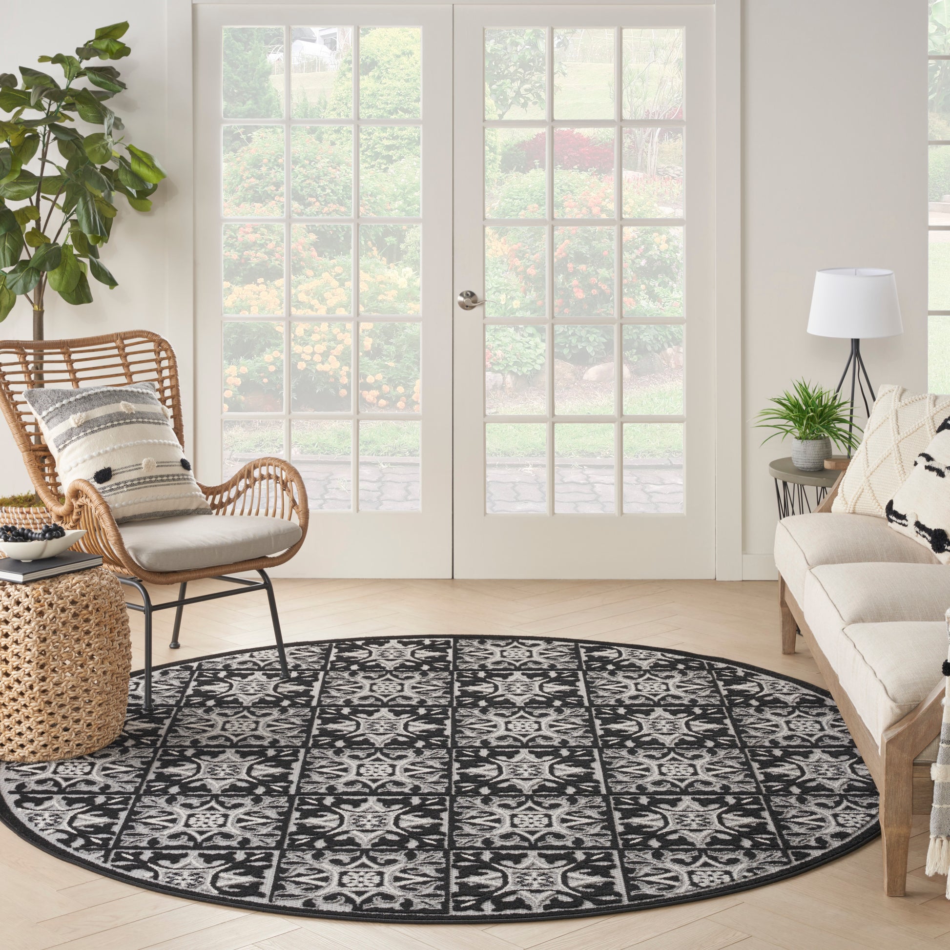 Round black-and-white contemporary patterned rug in a bright living room with wicker chair, beige sofa, plants and French doors.