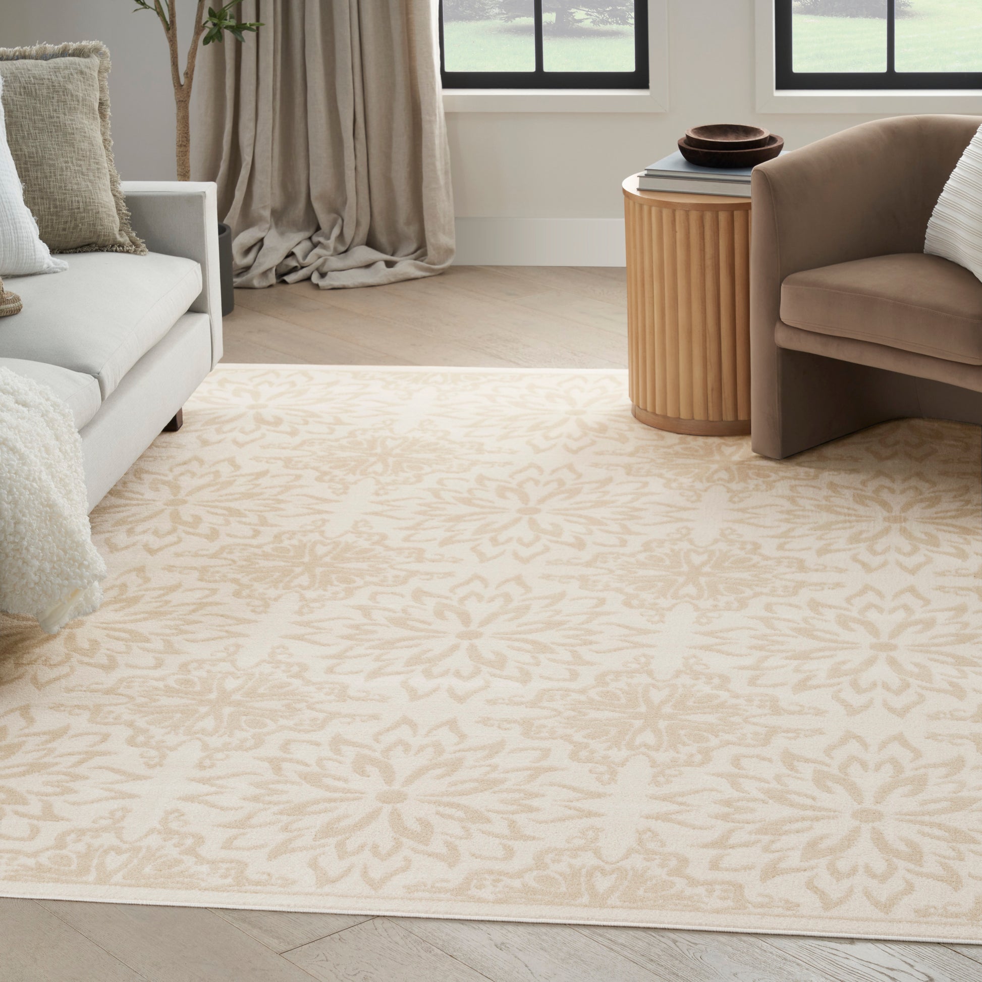 Beige floral-patterned area rug in a bright living room with a gray sofa, tan armchair, and a round ribbed wooden side table.