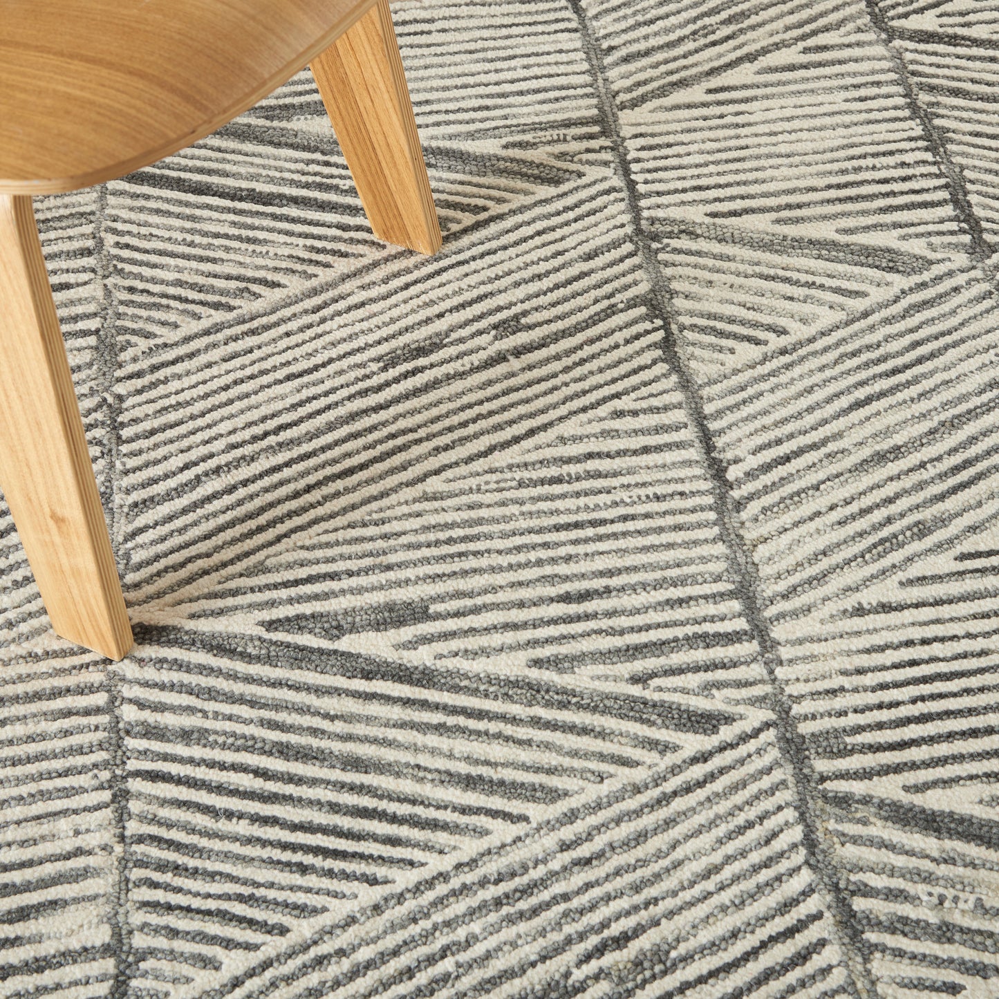 Light wooden chair leg with curved seat on a textured beige and gray geometric rug with diagonal stripe pattern.