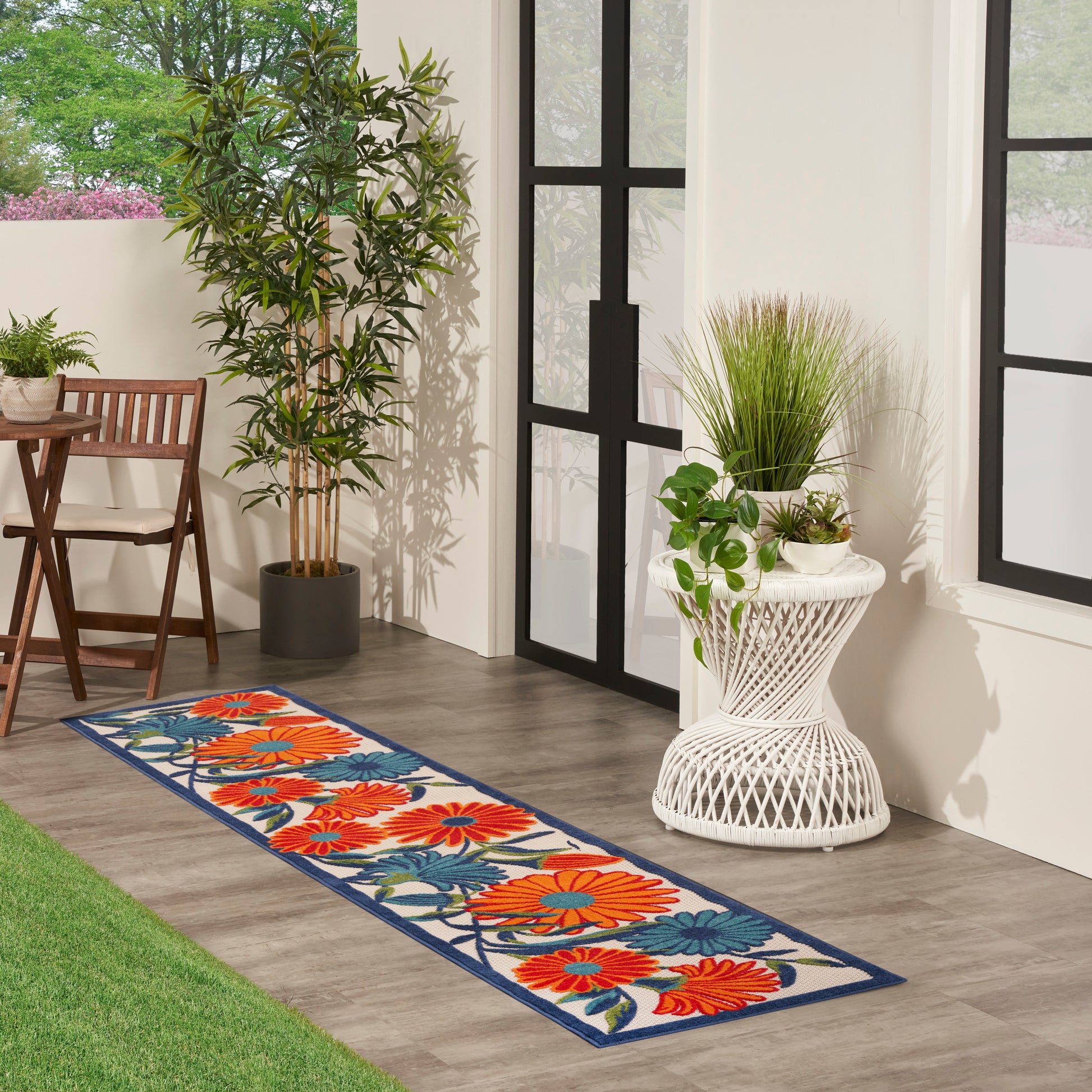 Floral runner rug with orange flowers, blue leaves and blue border on cream background beside glass doors, with potted plants.