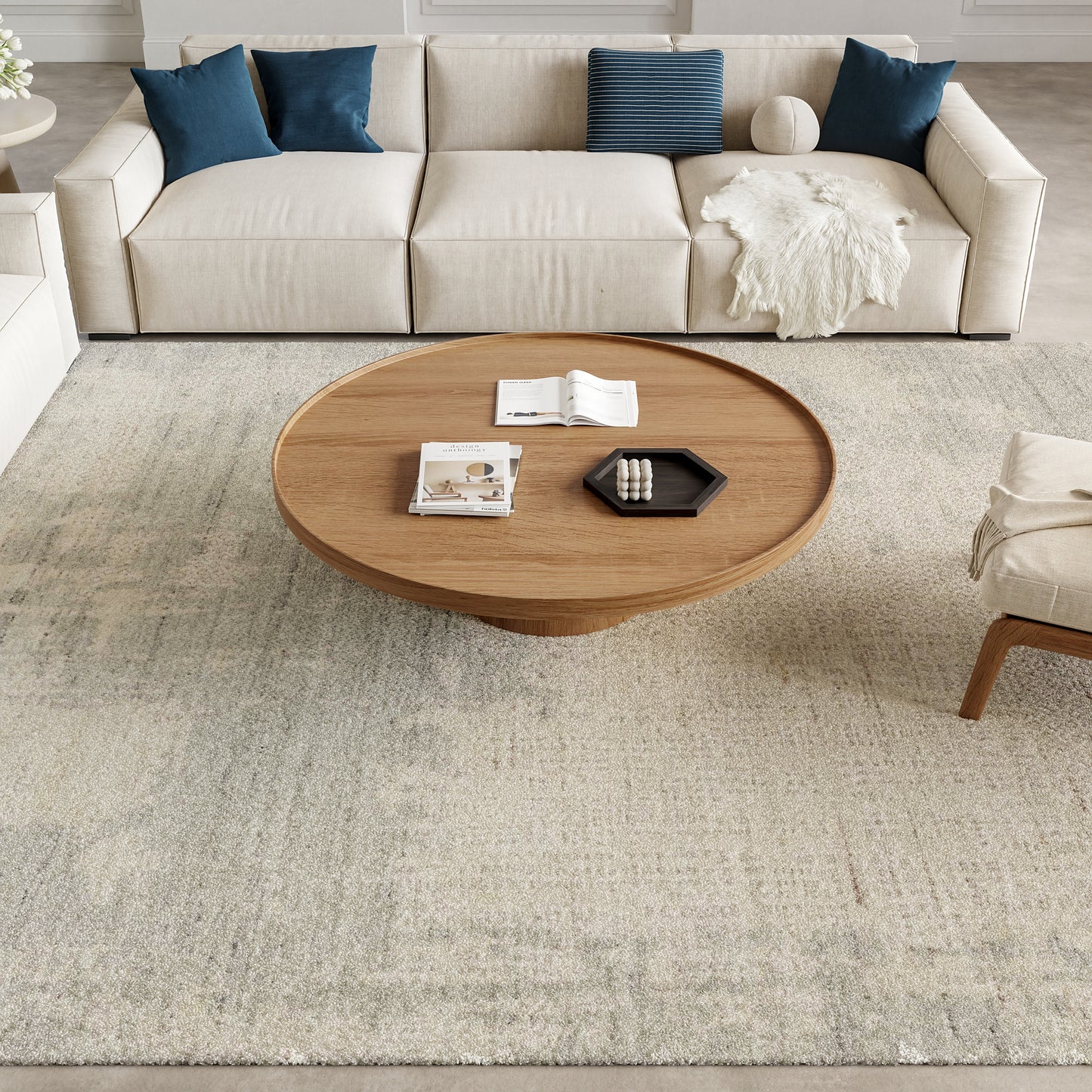 Round wooden coffee table with pedestal base, beige sectional sofa with blue cushions on a light textured rug.