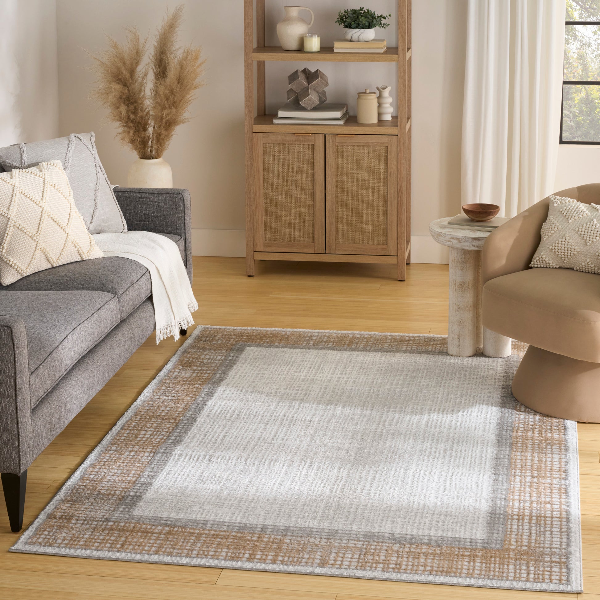 Neutral living room with gray sofa, beige chair, tan-border woven rug, and wooden cabinet with woven doors.