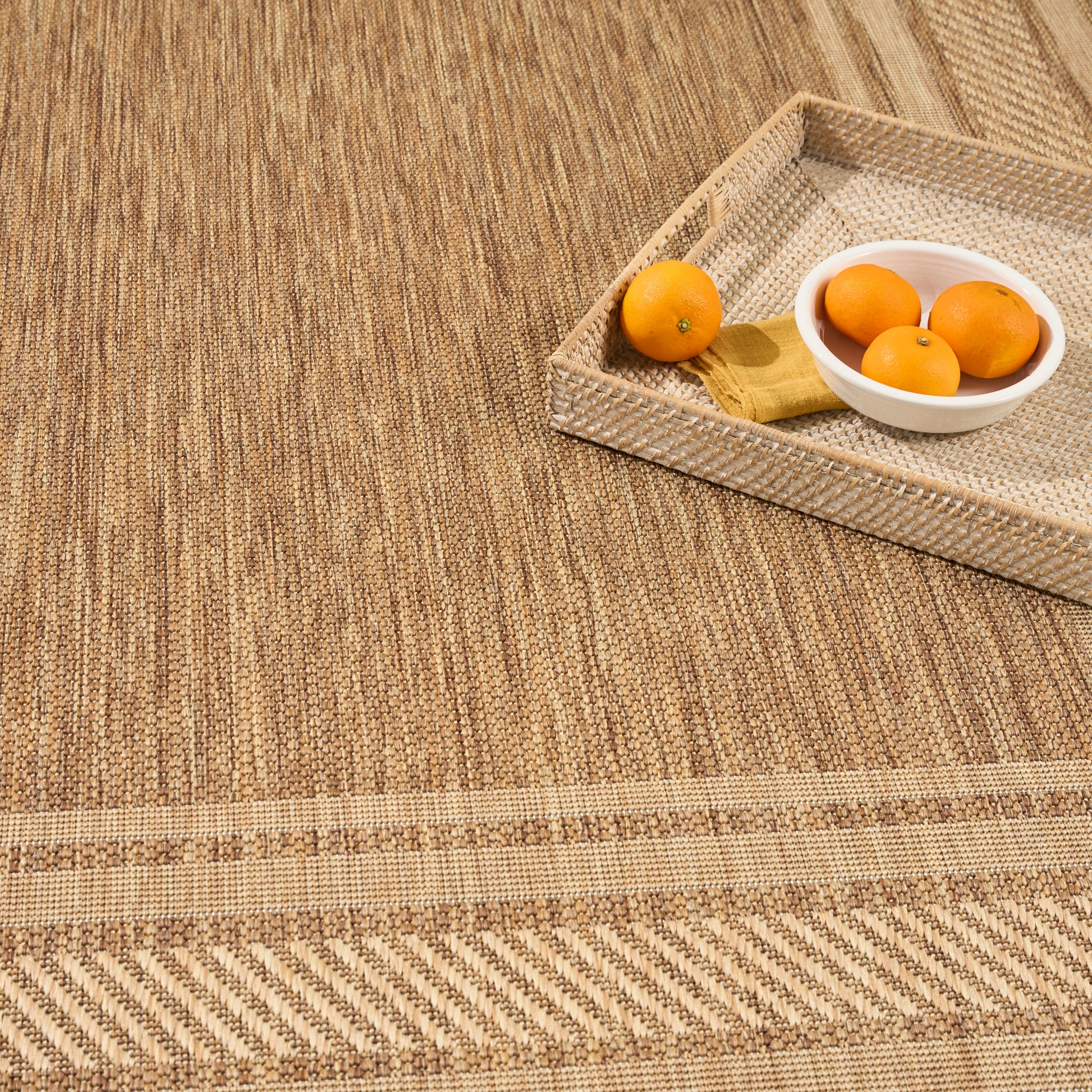 Beige woven mat with a rectangular wicker tray holding a white bowl of oranges and a loose orange beside a yellow cloth.