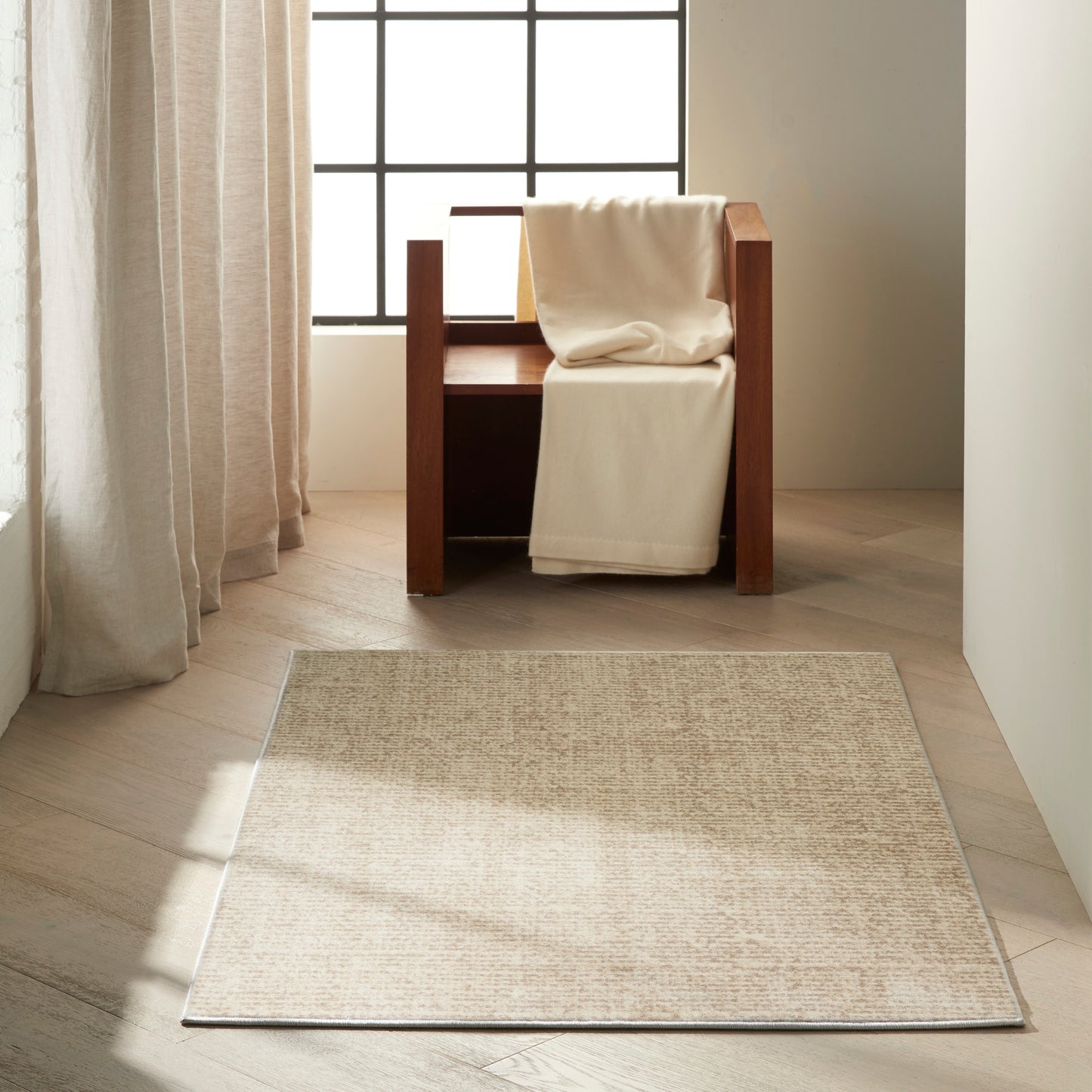 Beige textured 3' x 5' area rug on light wood floor beside a wooden armchair with a cream throw under a sunlit grid window.