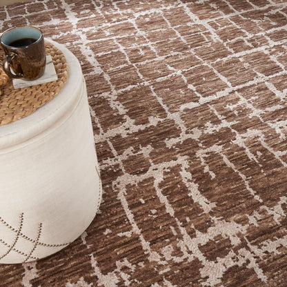 Beige fabric pouf with studded edge, round woven rattan tray holding a dark brown mug on a brown lattice rug.