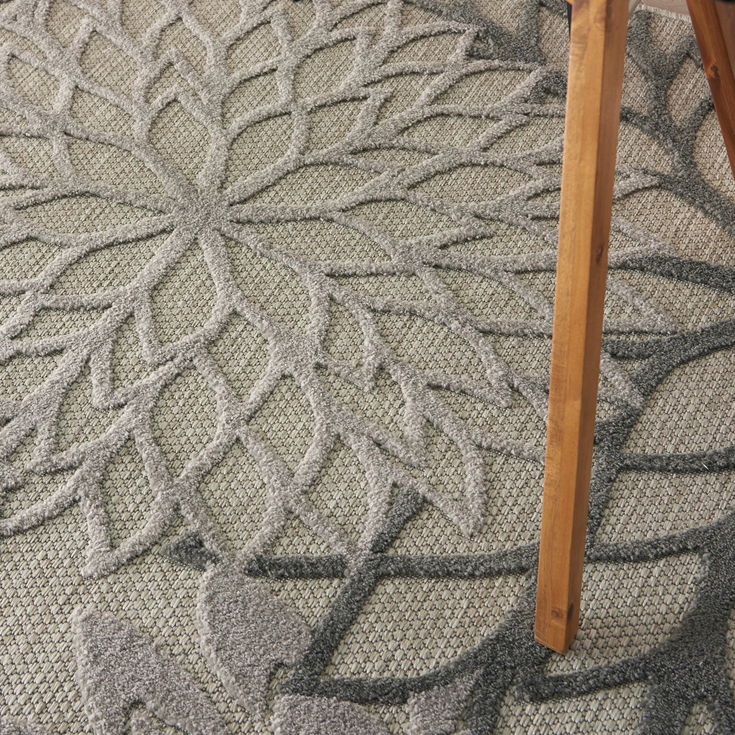 Close-up of a gray textured rug with a large branching leaf motif; wooden chair leg resting on the right.