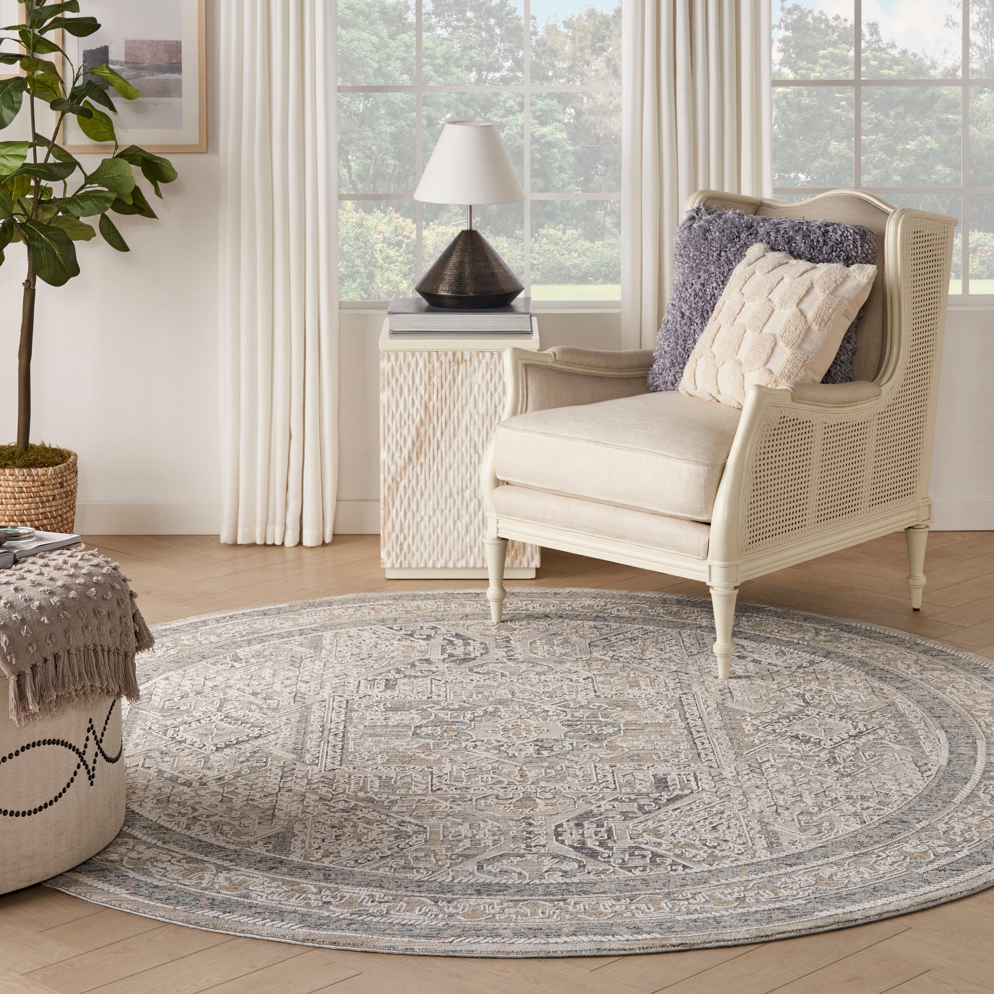 Round gray-blue patterned rug under cream wicker armchair with beige cushion and lilac throw by window.