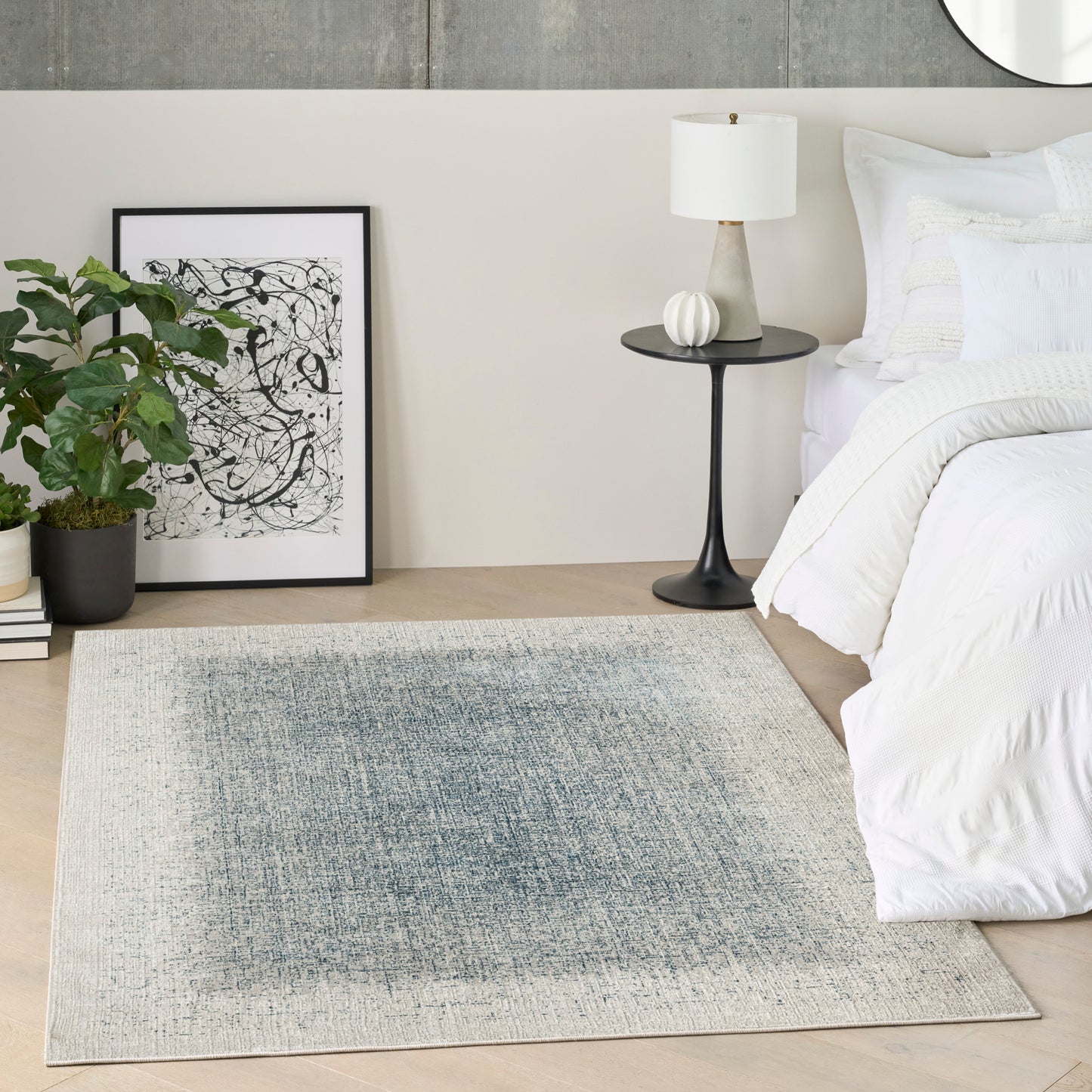 Neutral bedroom with white bedding, black bedside table and lamp, framed abstract art, potted plant, and blue-gray rug.