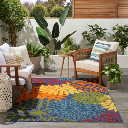 Bold tropical-leaf outdoor rug in blue, orange, yellow, and green on a gray deck with wooden chairs and lush potted plants.