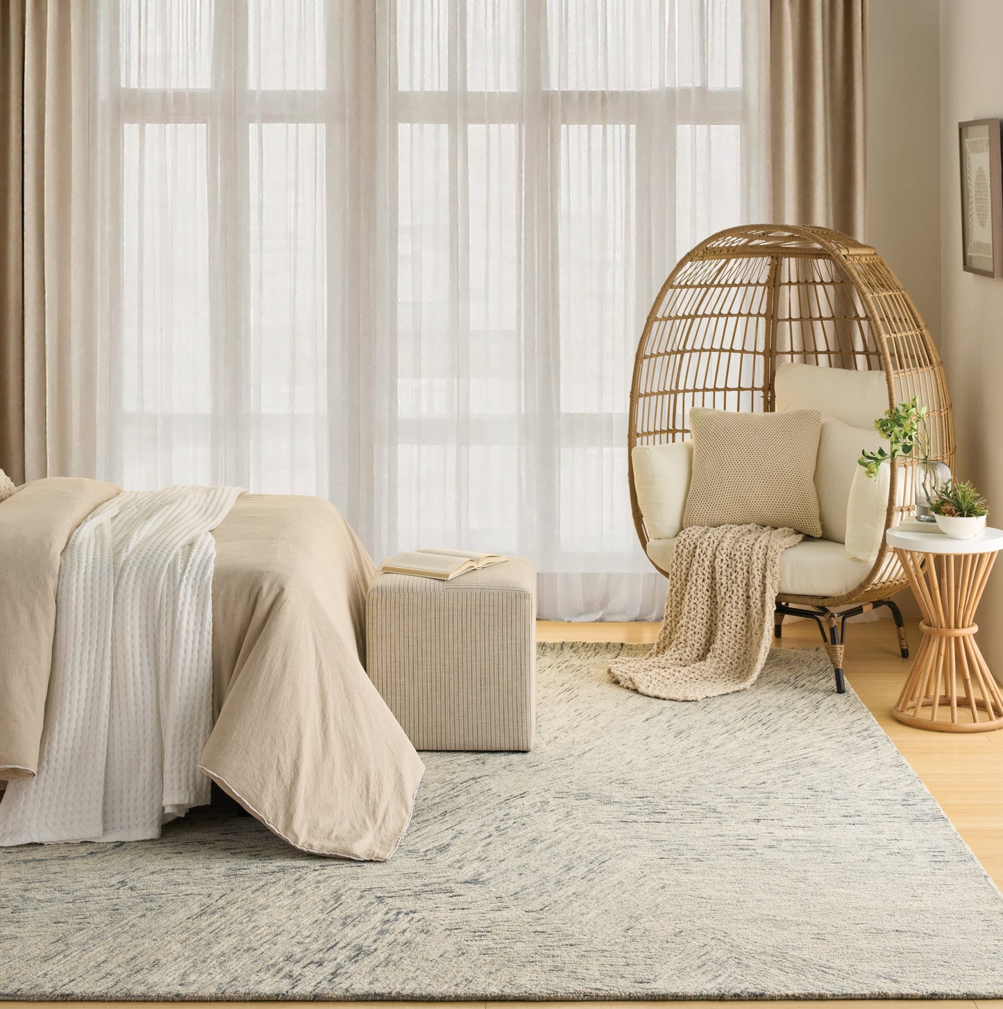 Neutral-toned bedroom with beige bedding, knitted throw, round rattan egg chair, ottoman, and a soft textured rug.