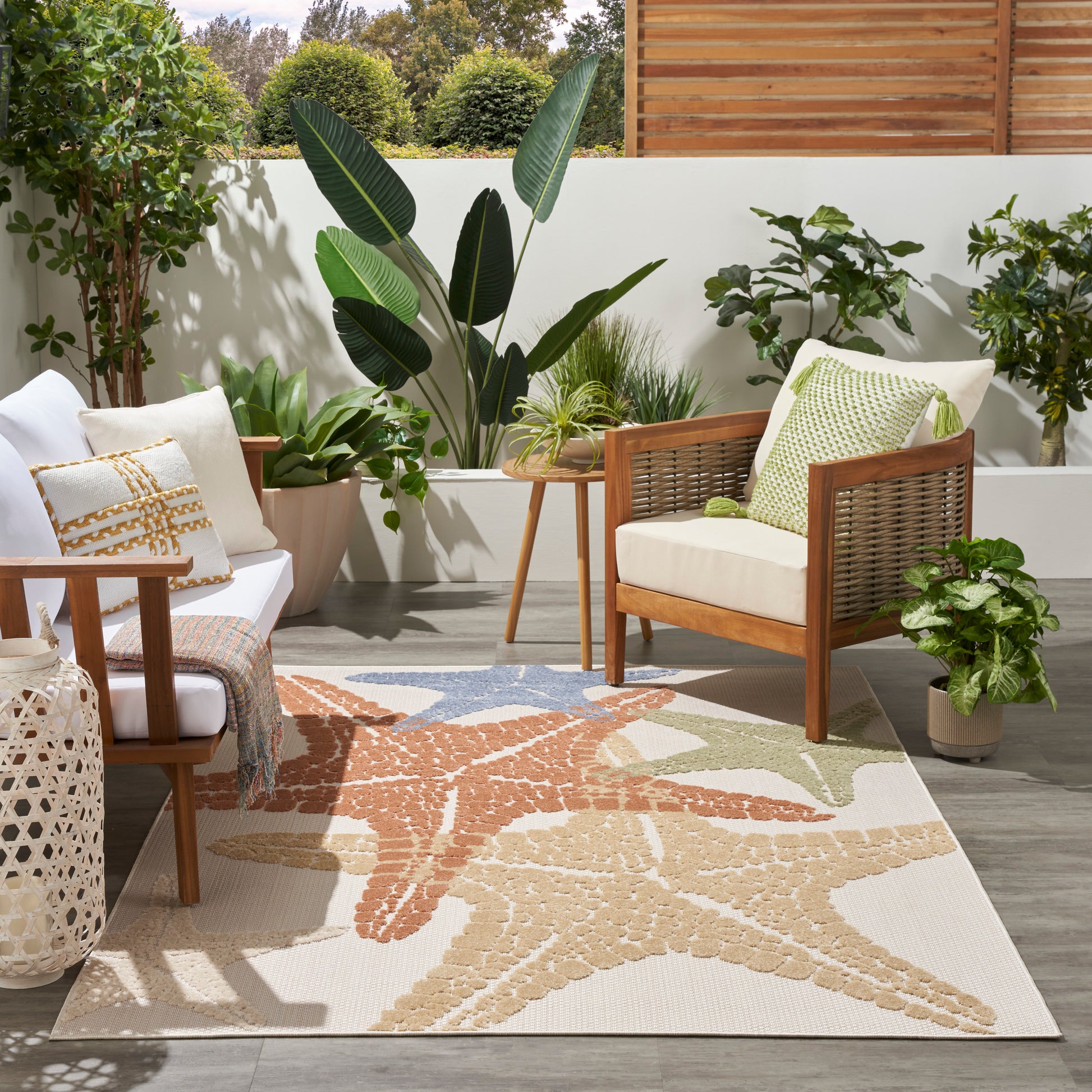 Outdoor patio seating with wooden frame chairs, white cushions, green pillow, lush potted plants, beige starfish-pattern rug.