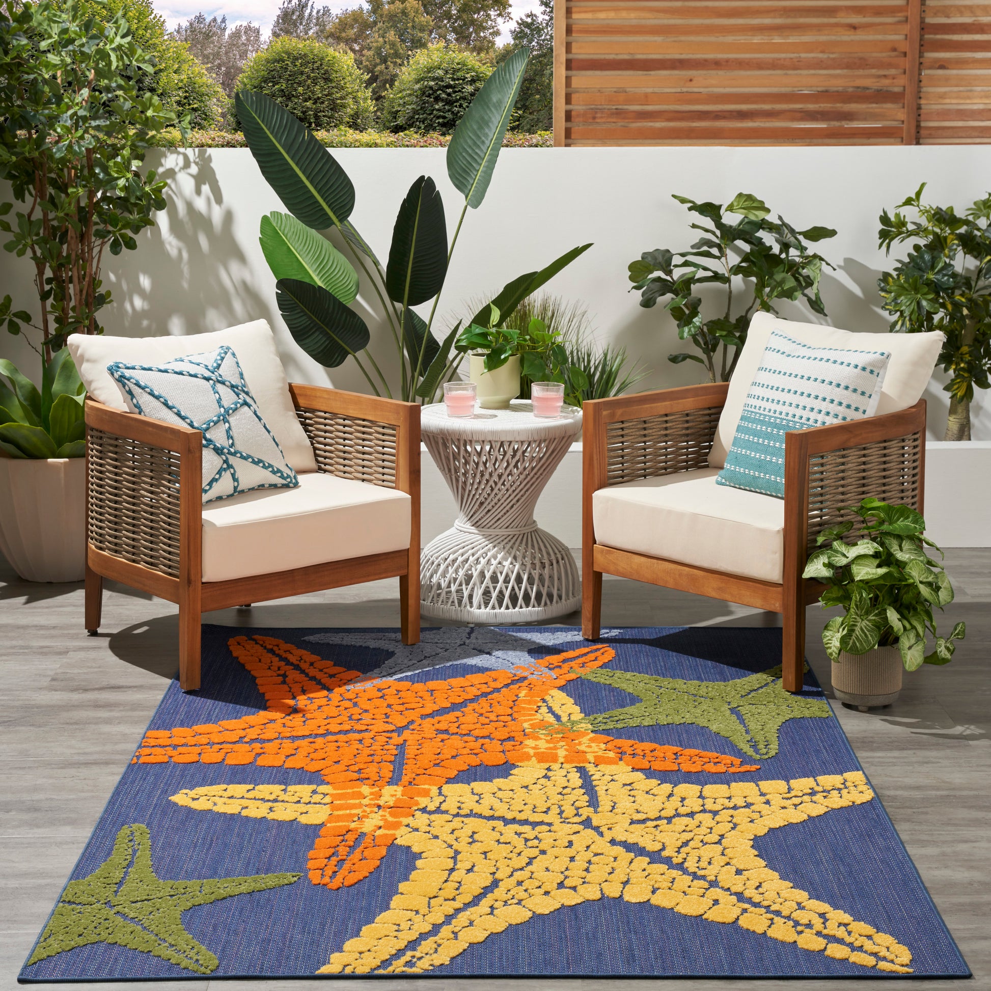 Outdoor patio with wicker-framed chairs, white cushions, blue starfish rug, tropical plants, and a white round table.
