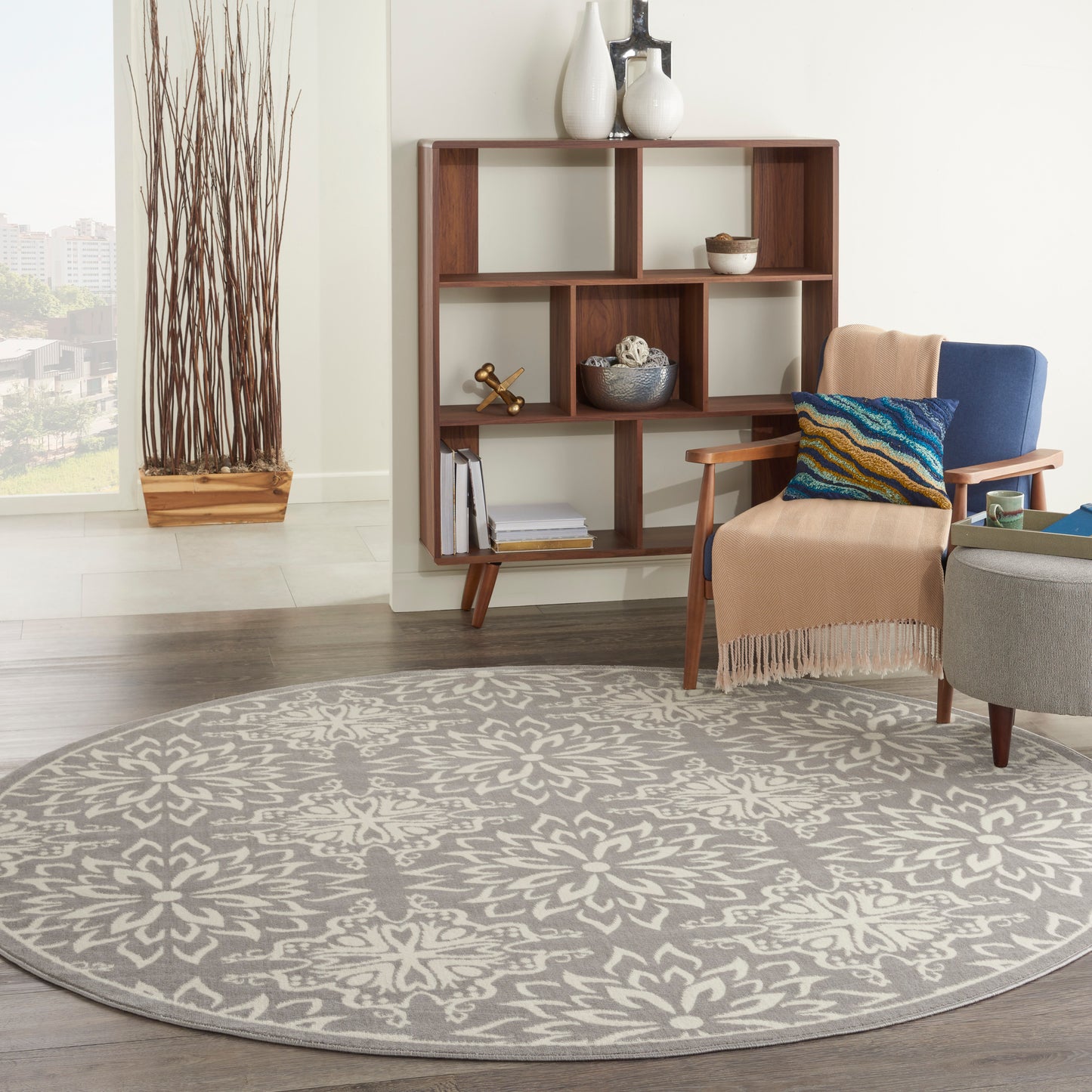 Living room with round grey patterned rug, mid-century shelf, beige armchair with blue pillow, ottoman, and tall twig vase.