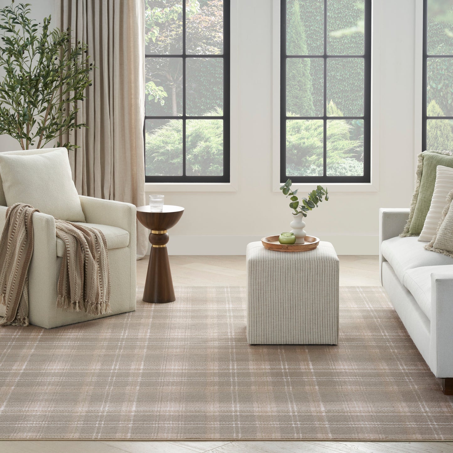 Neutral living room with cream sofa, beige armchair, plaid rug, black-framed windows, wooden table, ribbed ottoman, greenery.