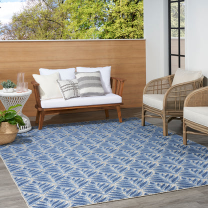 Blue leaf-patterned outdoor rug beneath white-cushioned wooden loveseat and wicker chairs on sunny balcony.