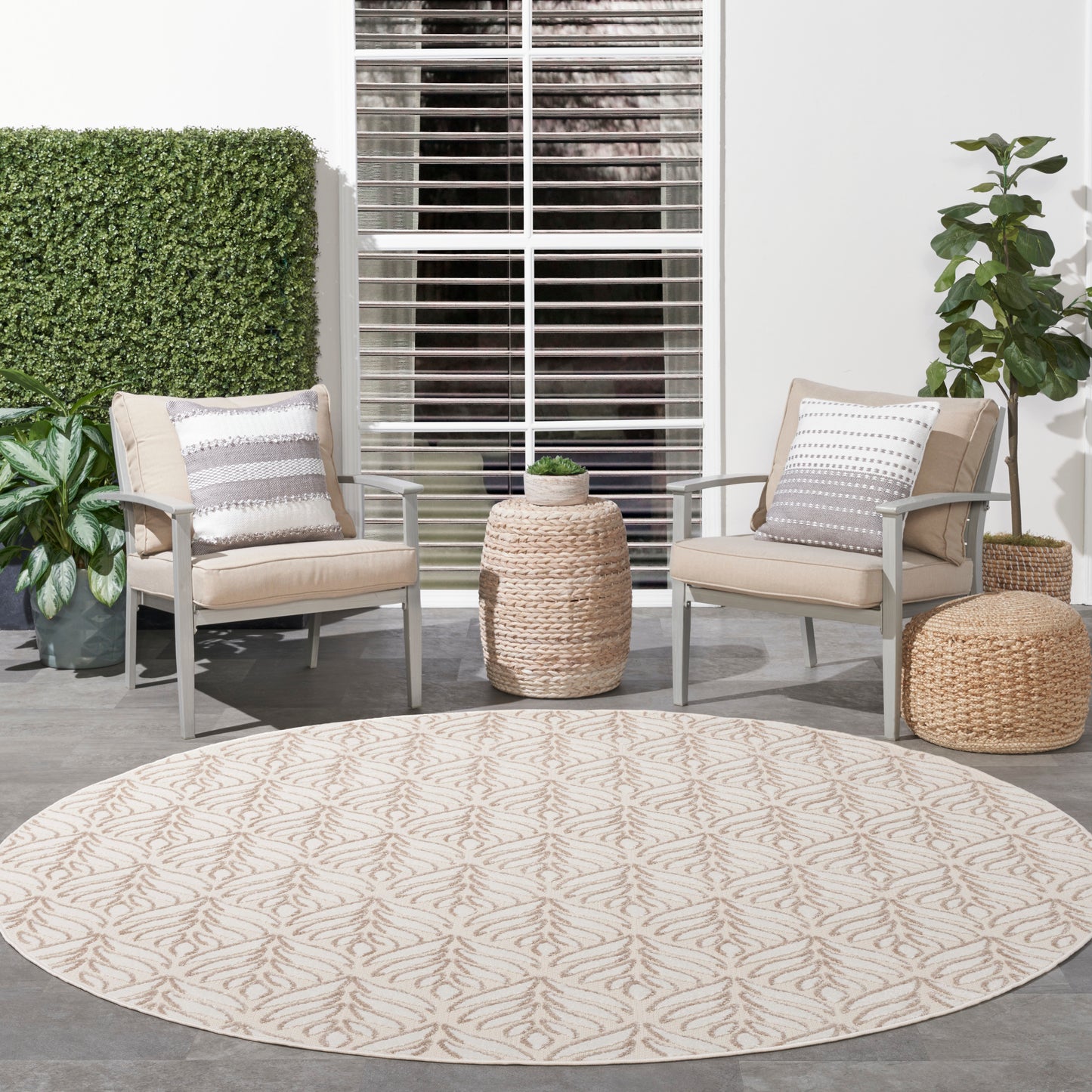 Round beige outdoor rug with subtle leaf pattern; two beige cushioned patio chairs, wicker tables, and potted plants.