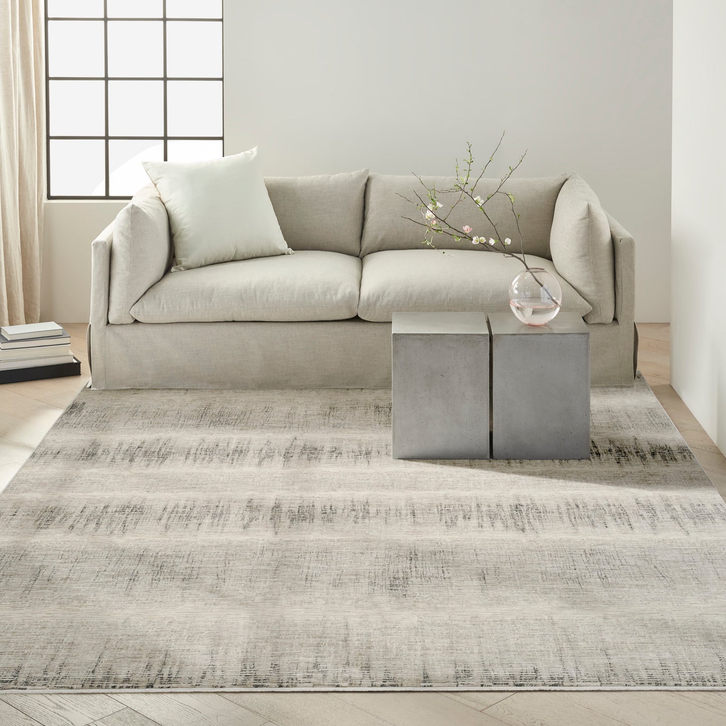 Neutral living room: beige sofa, light gray rug, two concrete block side tables, glass vase with flowering branches.