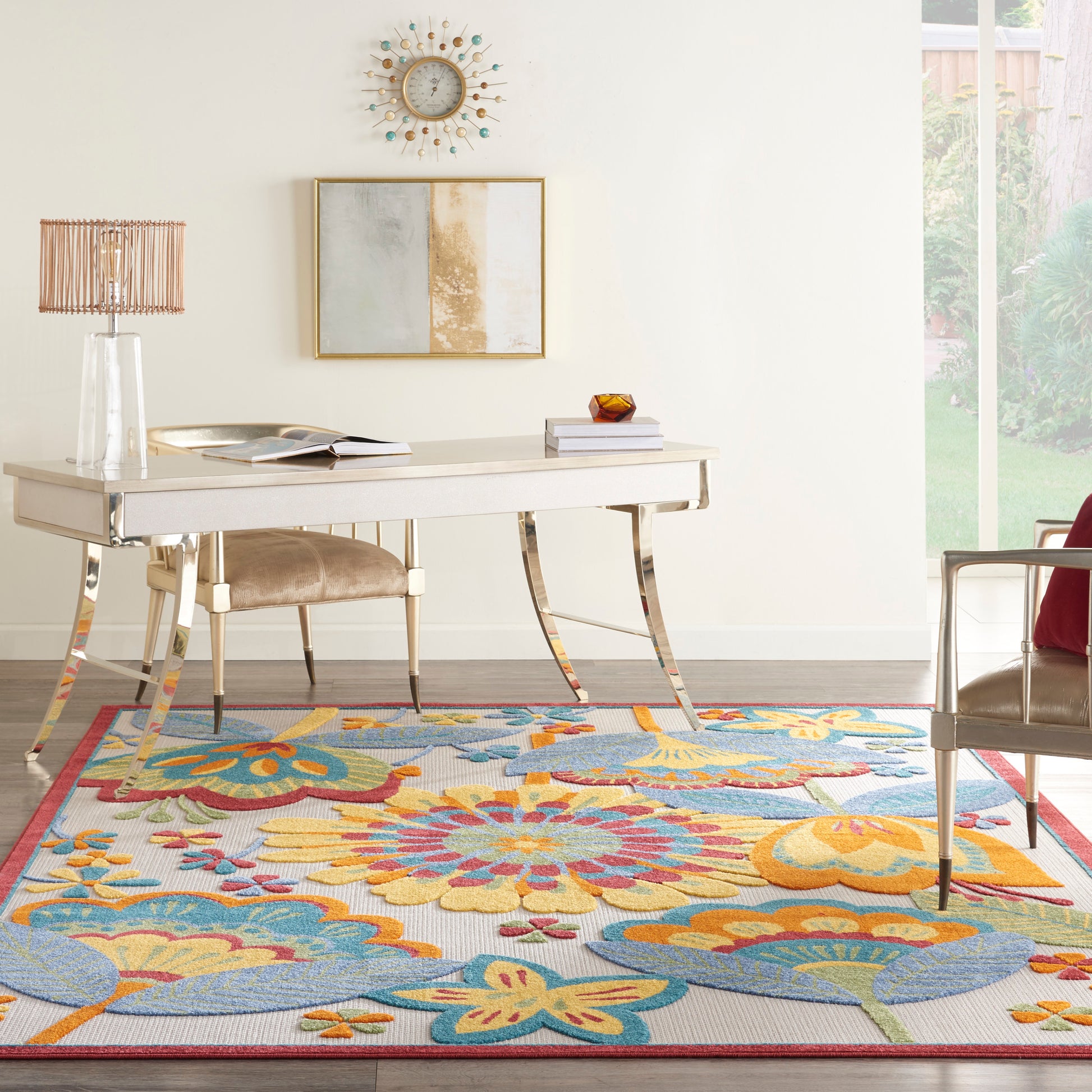 White desk with brass curved legs, glass table lamp, beige upholstered chair, & a colorful floral rug with blues, yellows.
