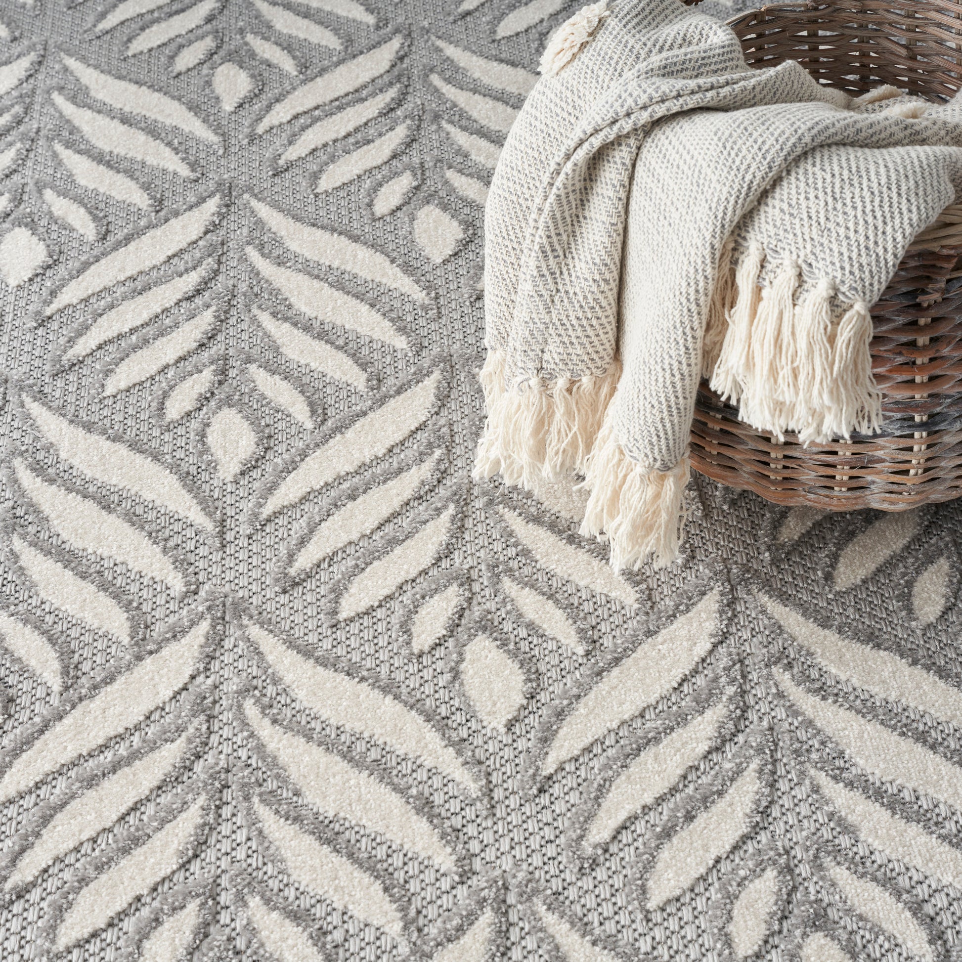 Grey and cream leaf-pattern rug with a cream fringed knit throw draped over a wicker basket.