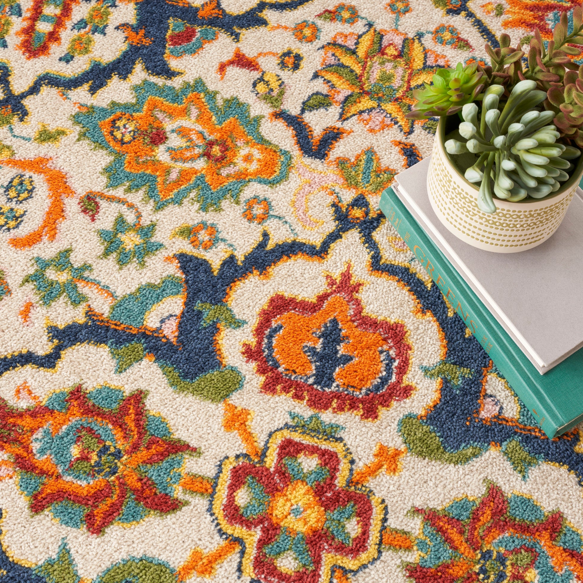 Colorful floral-patterned rug with cream background; beige planter with succulents atop stacked teal and gray books.