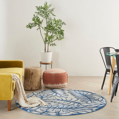Bright living room corner: yellow sofa, round blue leaf rug, wooden stools, potted tree, cream throw, black metal chair.