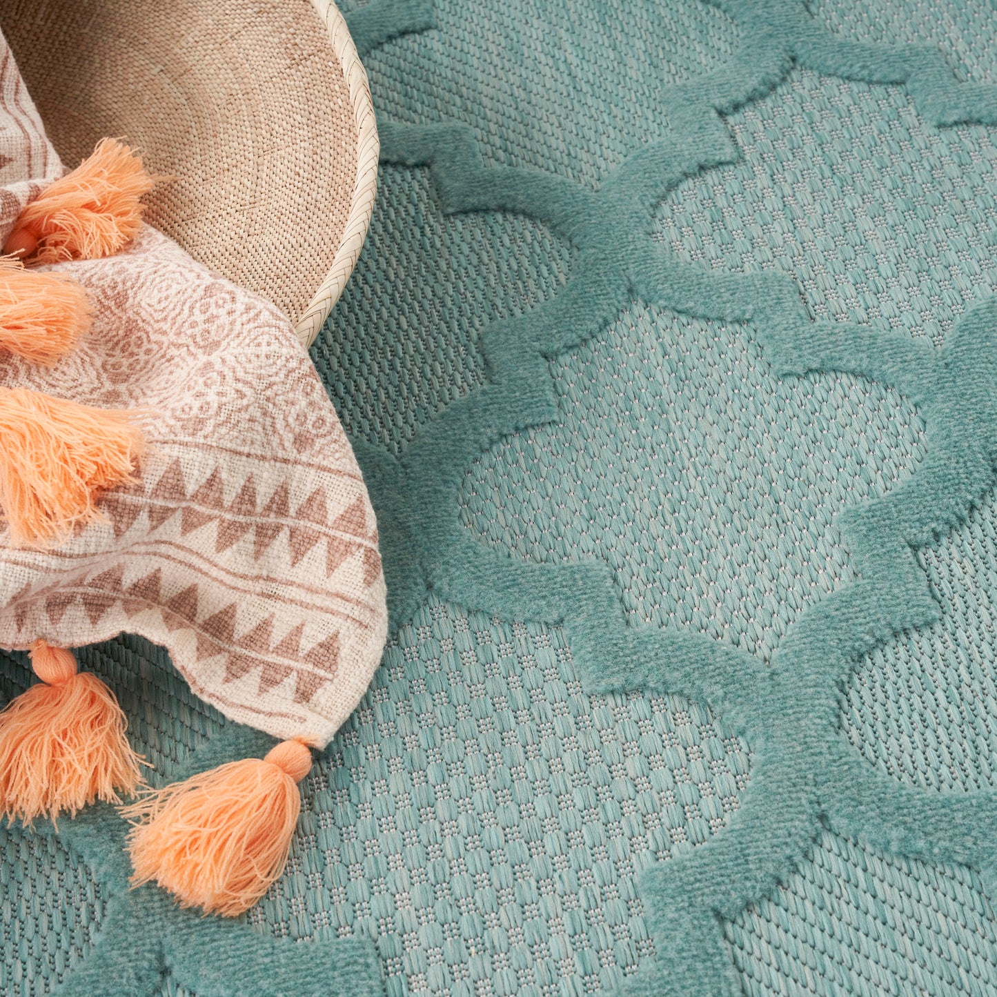 Teal textured rug with lattice/trellis pattern; beige tasseled throw with orange tassels draped on edge.