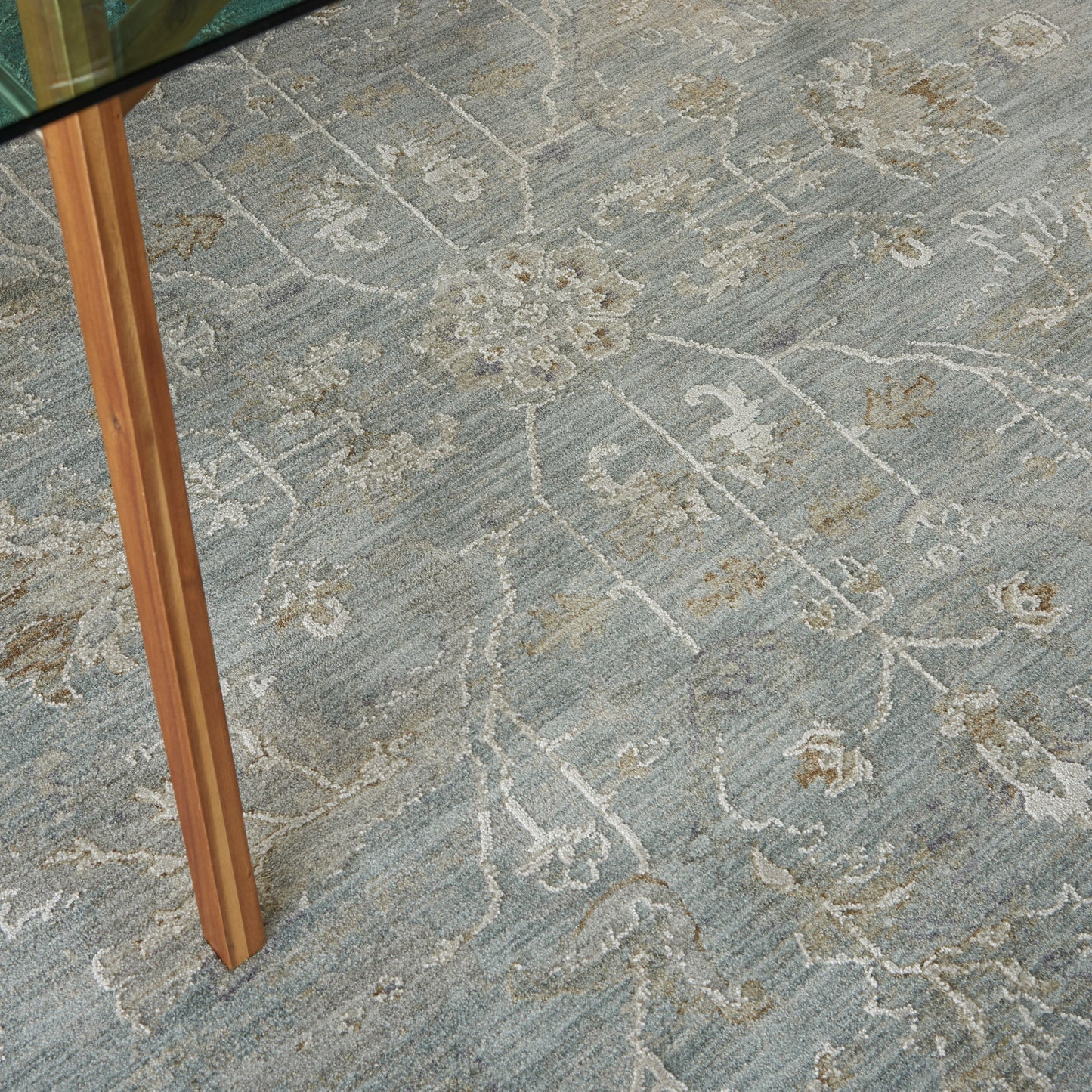 Blue-gray area rug with cream floral vine pattern beneath a glass-top wooden table.