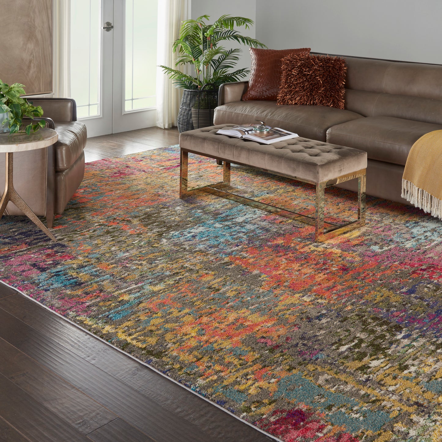 Colorful abstract area rug in a modern living room with a brown leather sofa, tufted ottoman, gold legs, and potted plants.