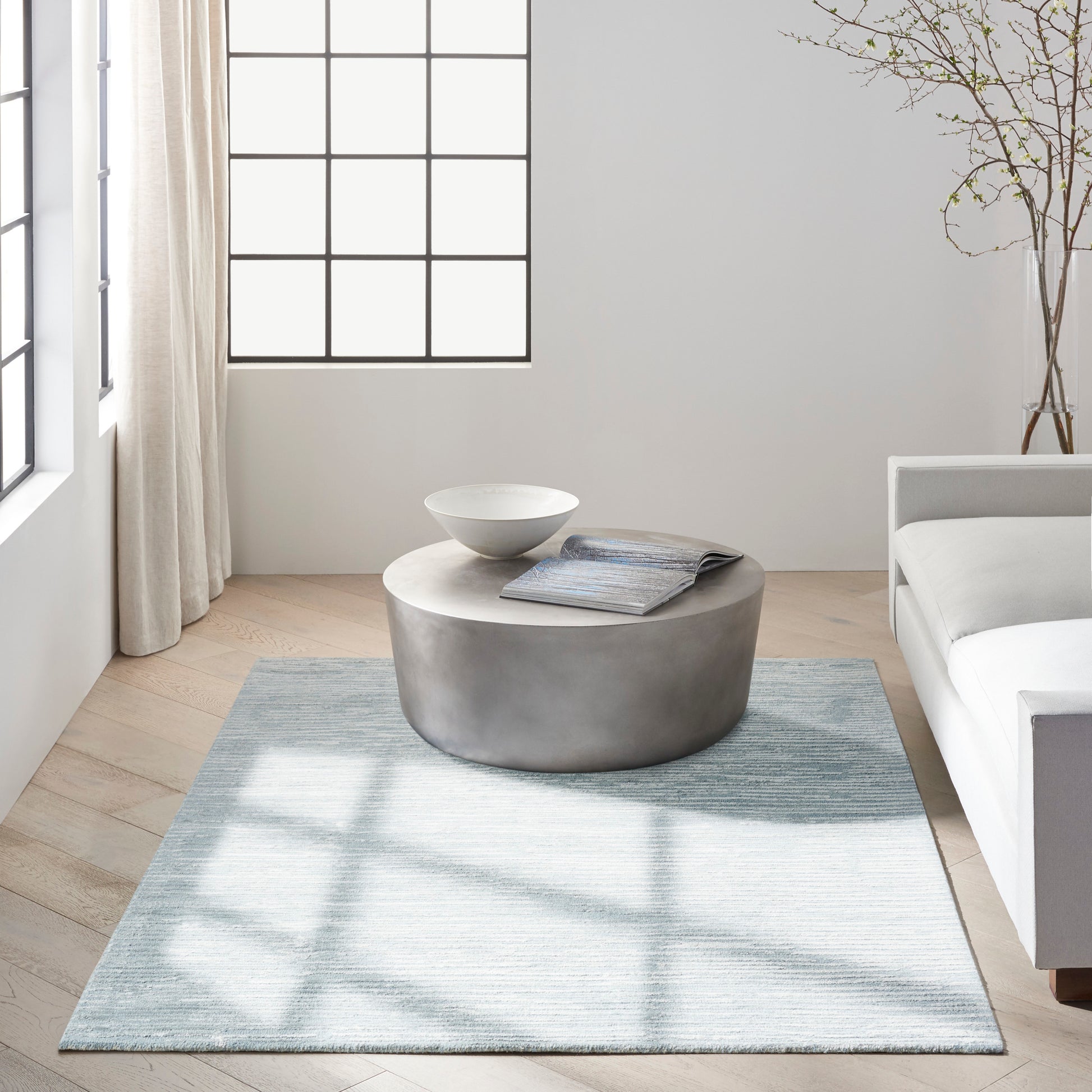Modern living room with a round metallic coffee table, white bowl, magazine, and a light blue rug beside a white sofa.