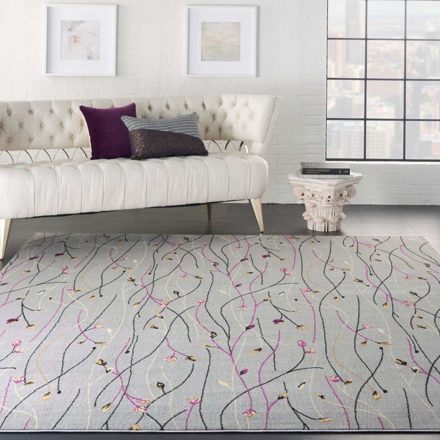 Living room with tufted white sofa, purple and gray cushions, city-view window, and gray rug with pink, gold, black vine swirls.