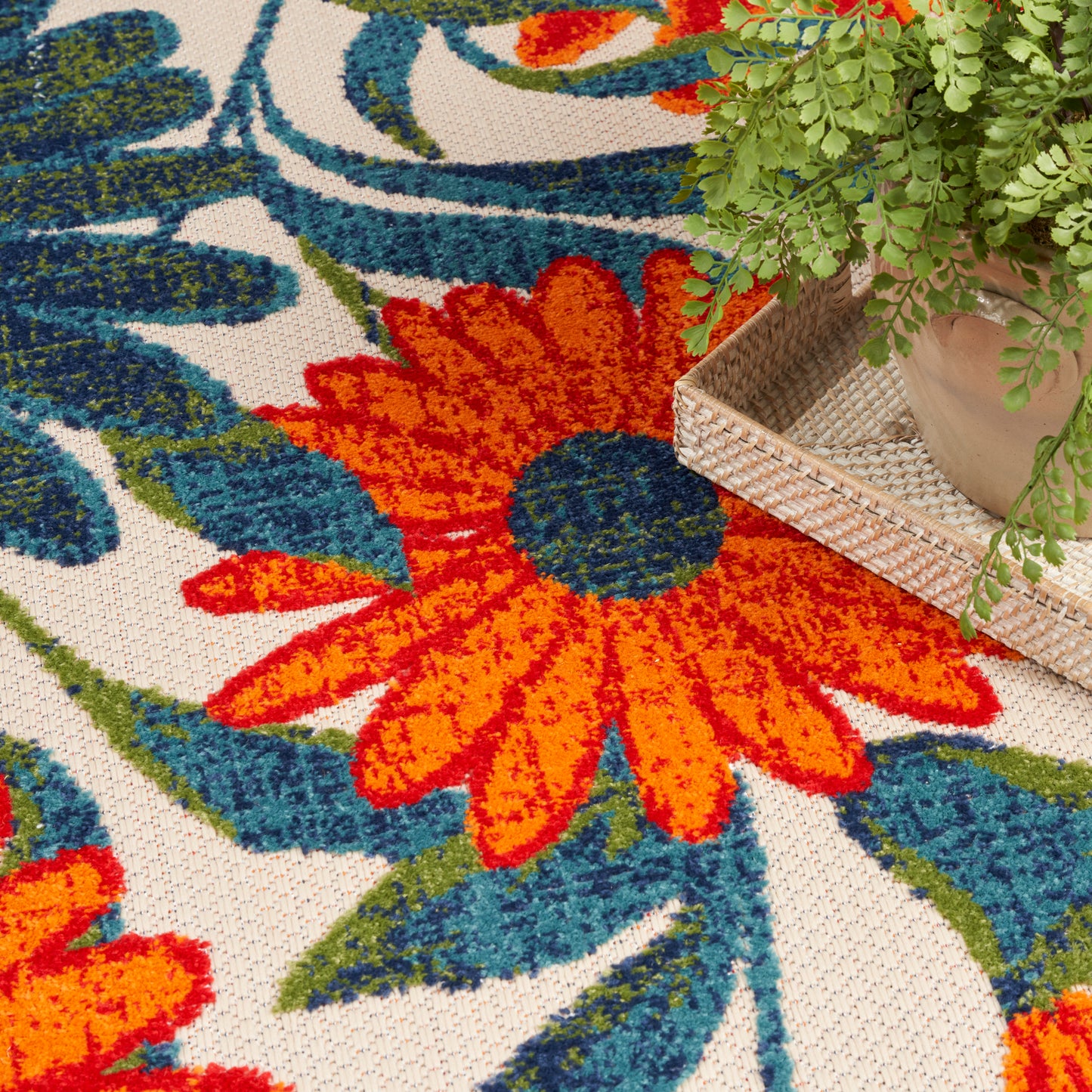 Vibrant floral outdoor rug with orange petals, teal-blue center and green leaves; woven wicker tray holds a potted fern.