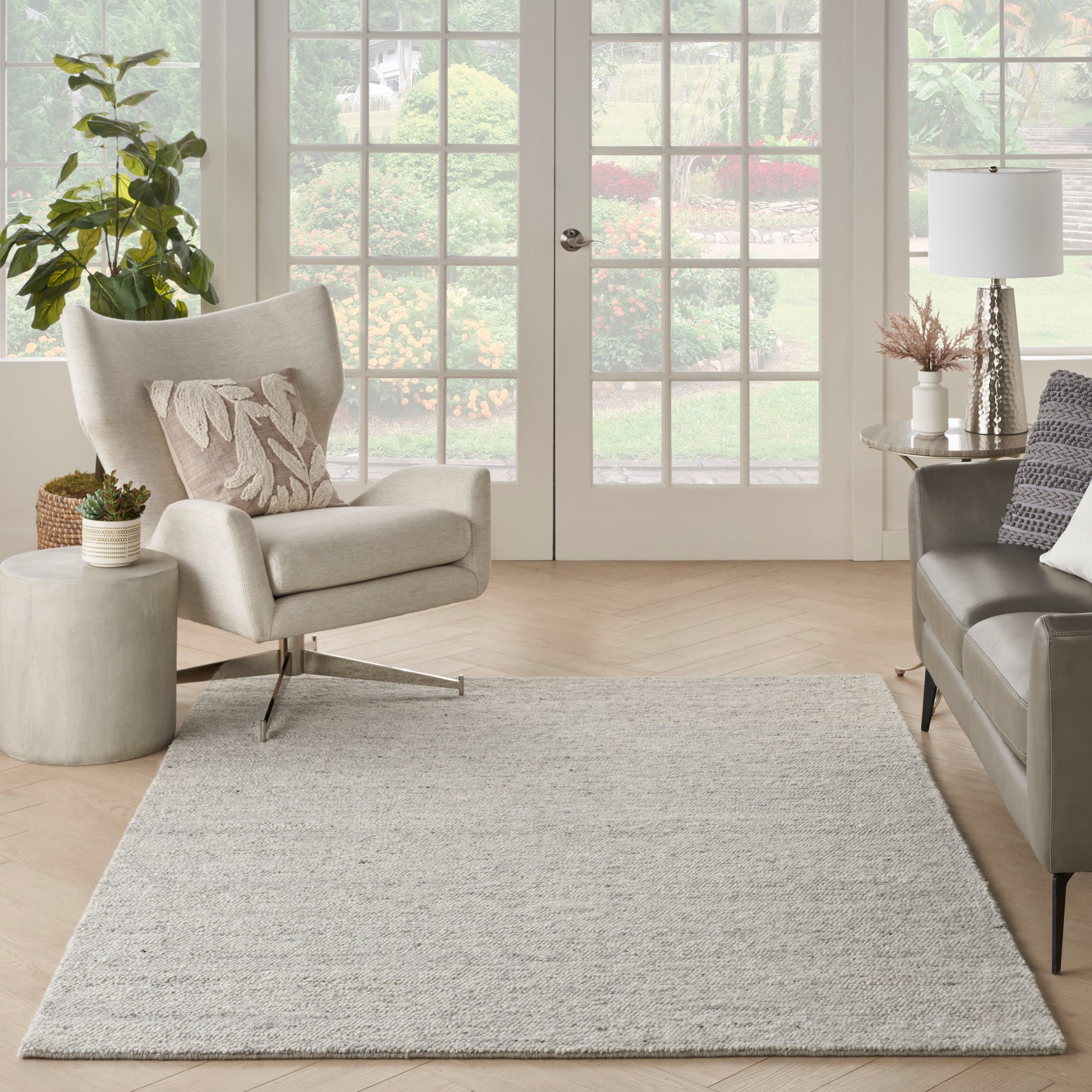 Light beige fabric armchair, gray sofa, textured neutral rug, potted plant, & white French doors in a bright living room.