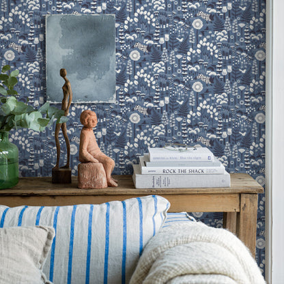 Wood console against blue floral wallpaper; green plant, two sculpted figurines, white books, and striped blue cushion.