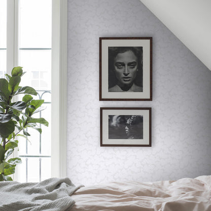 Tall green plant beside a bed with beige bedding, lavender wallpaper, and two dark wood-framed monochrome photos.